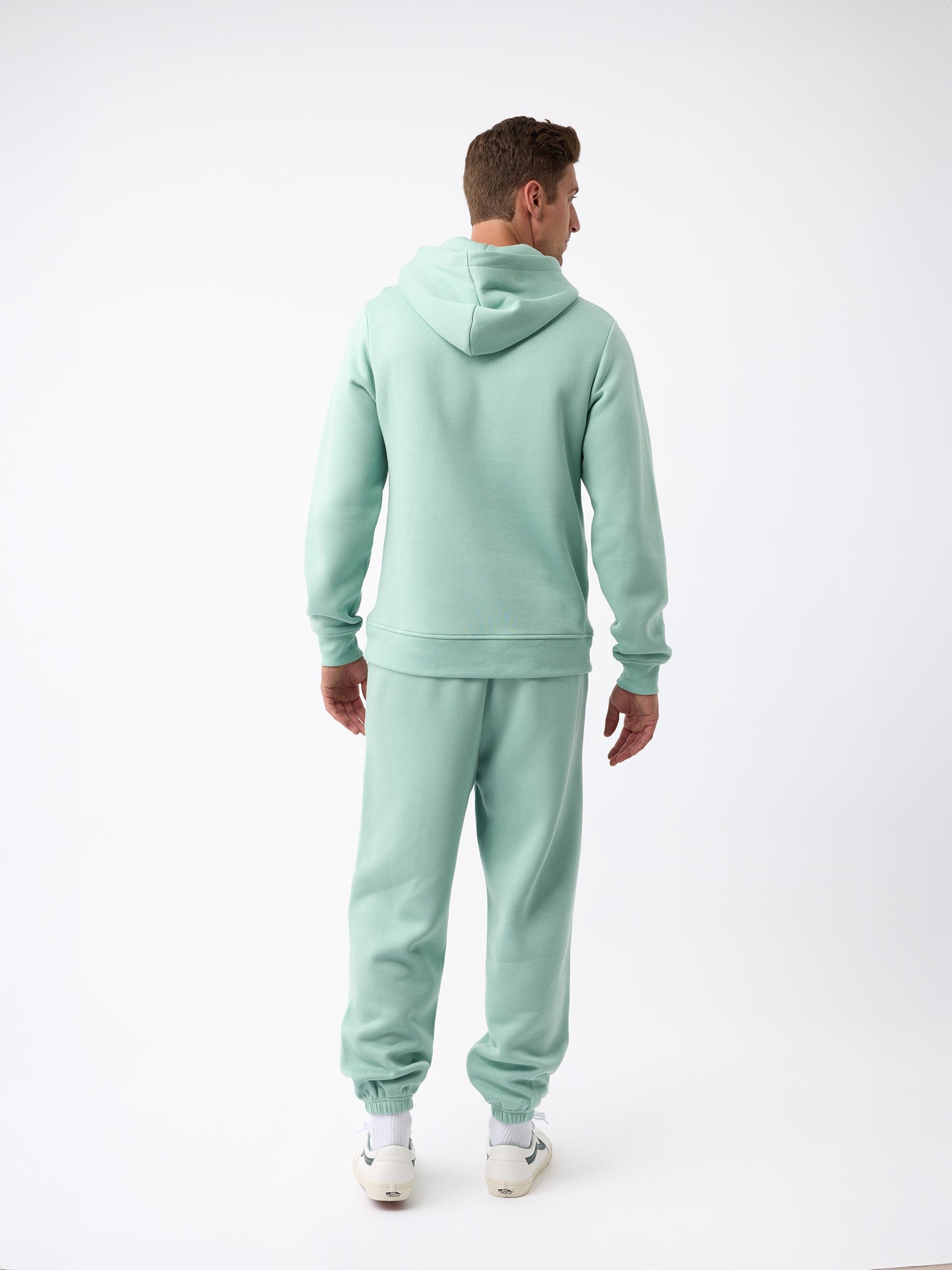 A person dressed in a Men's CityScape Sweatpant from Cozy Earth is facing away from the camera. They are standing against a plain white background, showcasing their light green hoodie and matching sweatpants, along with visible white sneakers. 