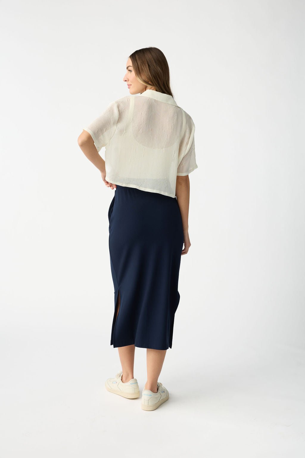 A person with long hair stands against a plain background, wearing a sheer white short-sleeve top and the navy blue Women's Brushed Bamboo Midi Skirt by Cozy Earth. They face away, hands by their sides and wear white sneakers. |Color:Navy