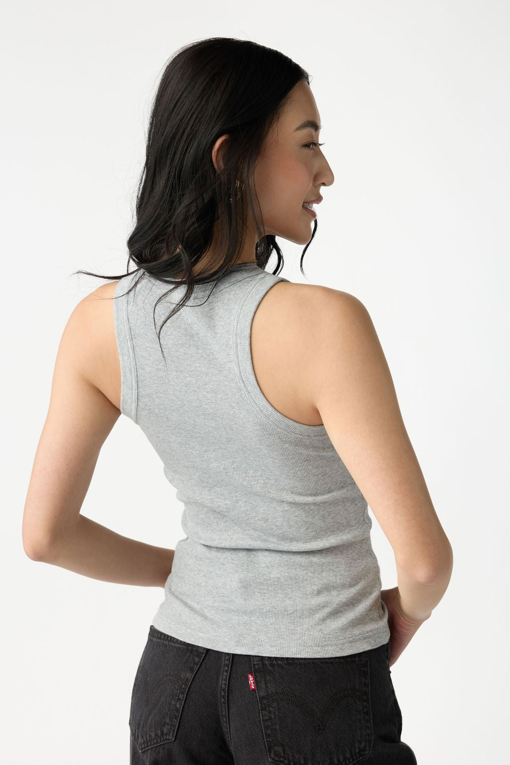 A dark haired woman wears Cozy Earth Women's Fine Ribbed Tank in Heather Grey. She faces away from the camera, turned to the side, and stands in front of a white background. 