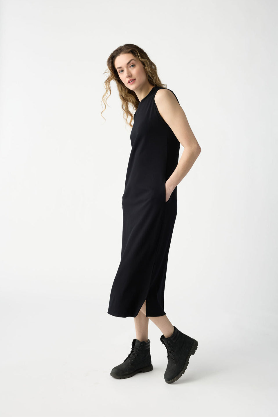 A person with long wavy hair is wearing Cozy Earth's Women's Brushed Bamboo Sleeveless Midi Dress and black boots. They stand sideways, hands in pockets, looking at the camera against a plain white backdrop. 