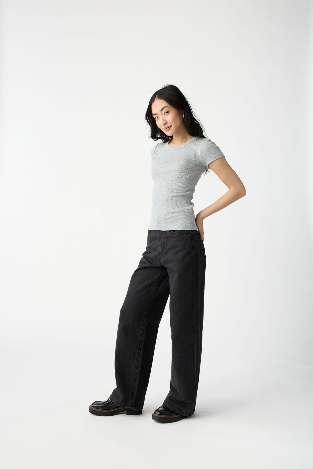 A dark haired woman wears Cozy Earth Women's Fine Ribbed Tee in Heather Grey. She is wearing black jeans and stands in front of a white background. 