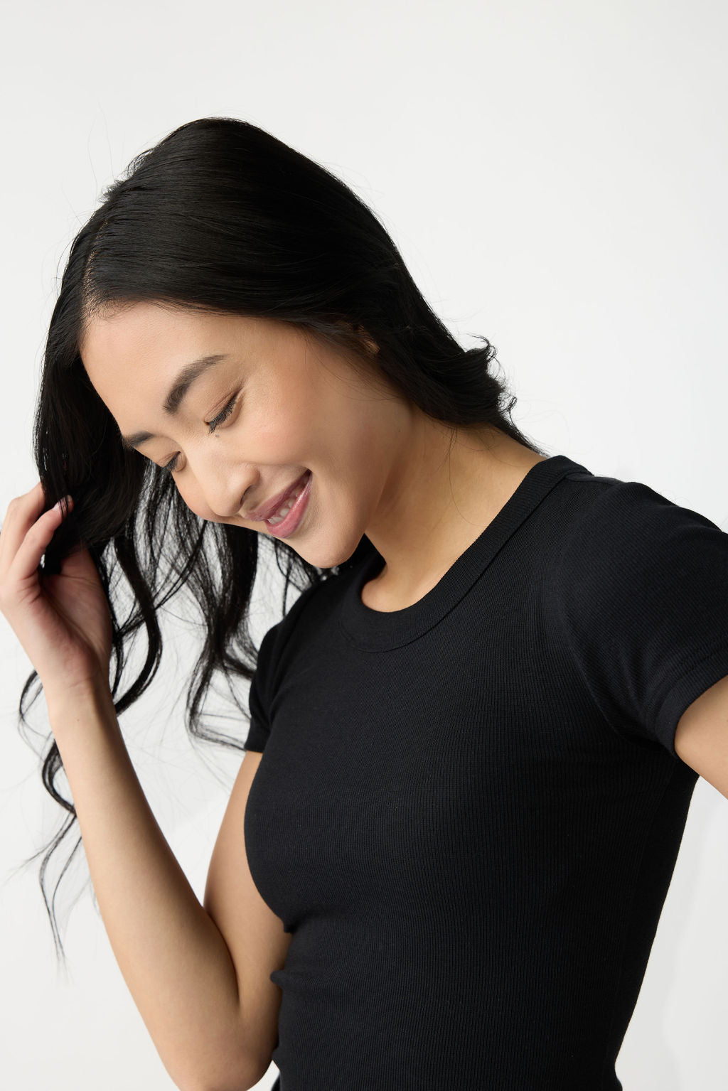 A dark haired woman wears Cozy Earth Women's Fine Ribbed Tee in Jet Black. She is pictured from the chest up and stands in front of a white background. 