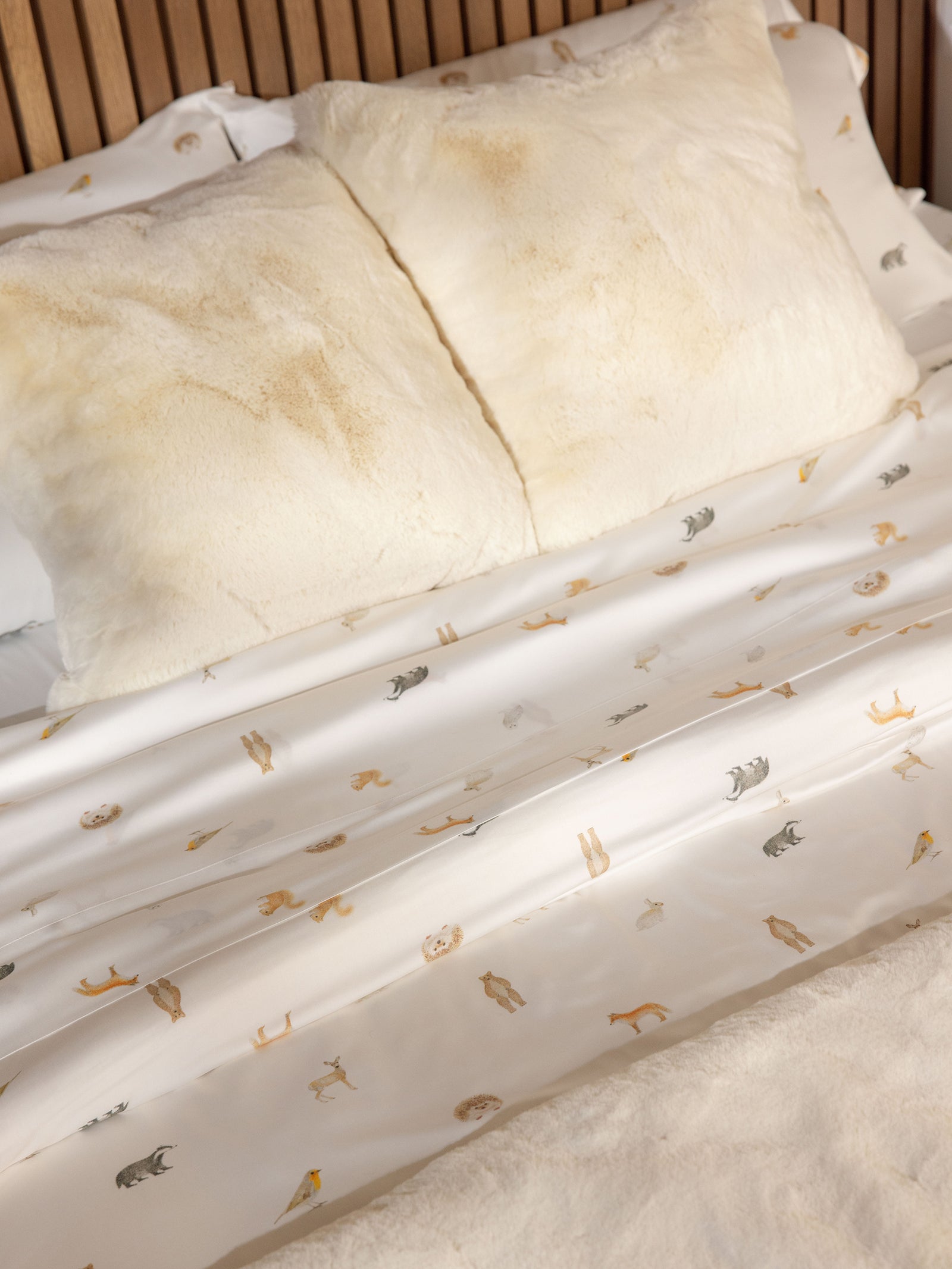 A neatly made bed with two large, cream-colored fluffy pillows draped with the Cozy Earth's Bamboo Duvet Cover. The subtle animal print of the sheets, showcasing silhouettes of various animals, creates a cozy and inviting atmosphere. The headboard features wooden vertical slats. 