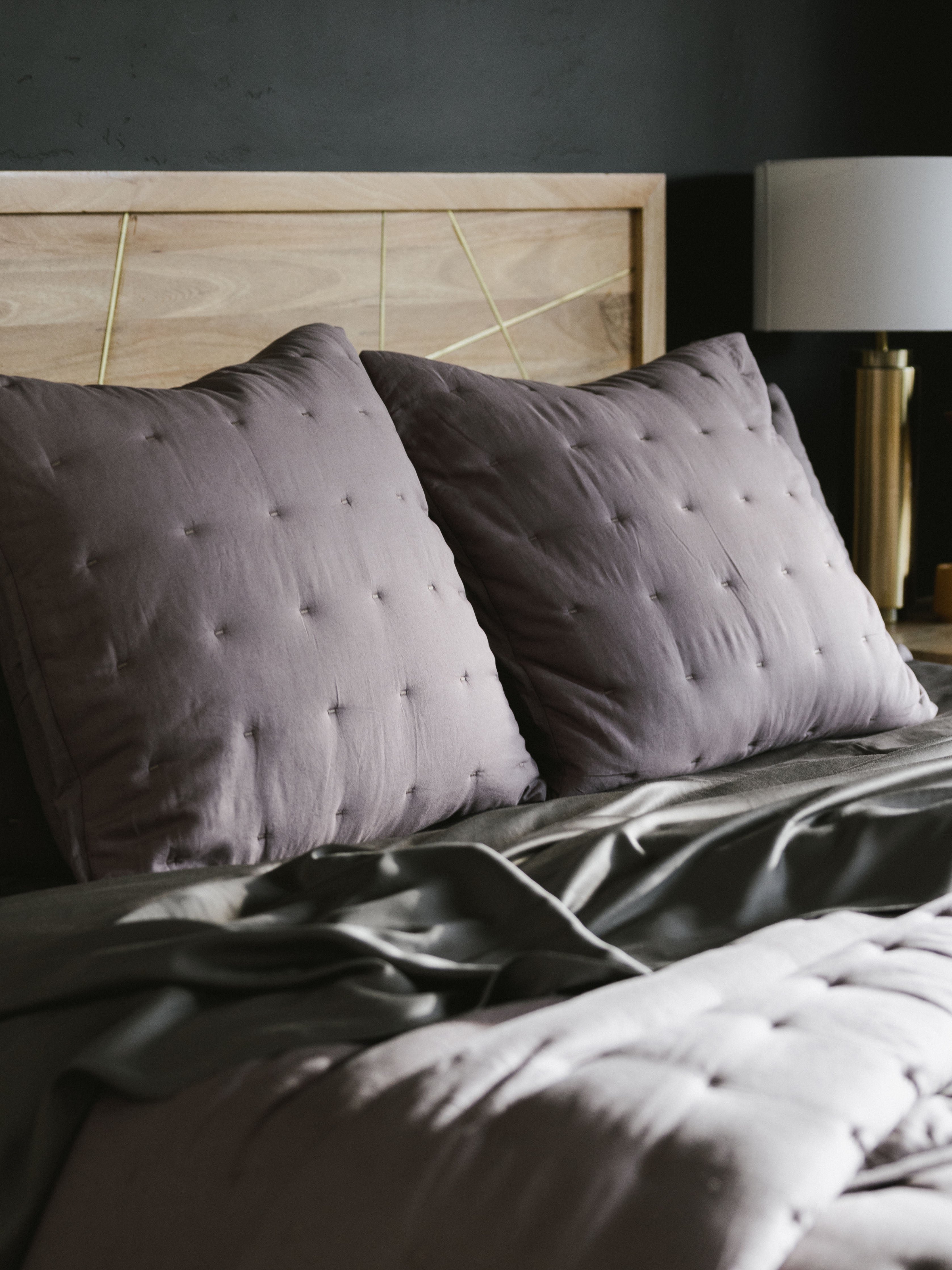 A cozy bed with Cozy Earth's Premium Down Alternative Insert covered in dark gray bedding and adorned with two large, tufted gray pillows rests against a light wood headboard. On the right side, a modern bedside lamp with a gold base adds elegance to the scene. The room's dark wall background creates a striking contrast with the light-colored headboard and pillows. |Size:Euro