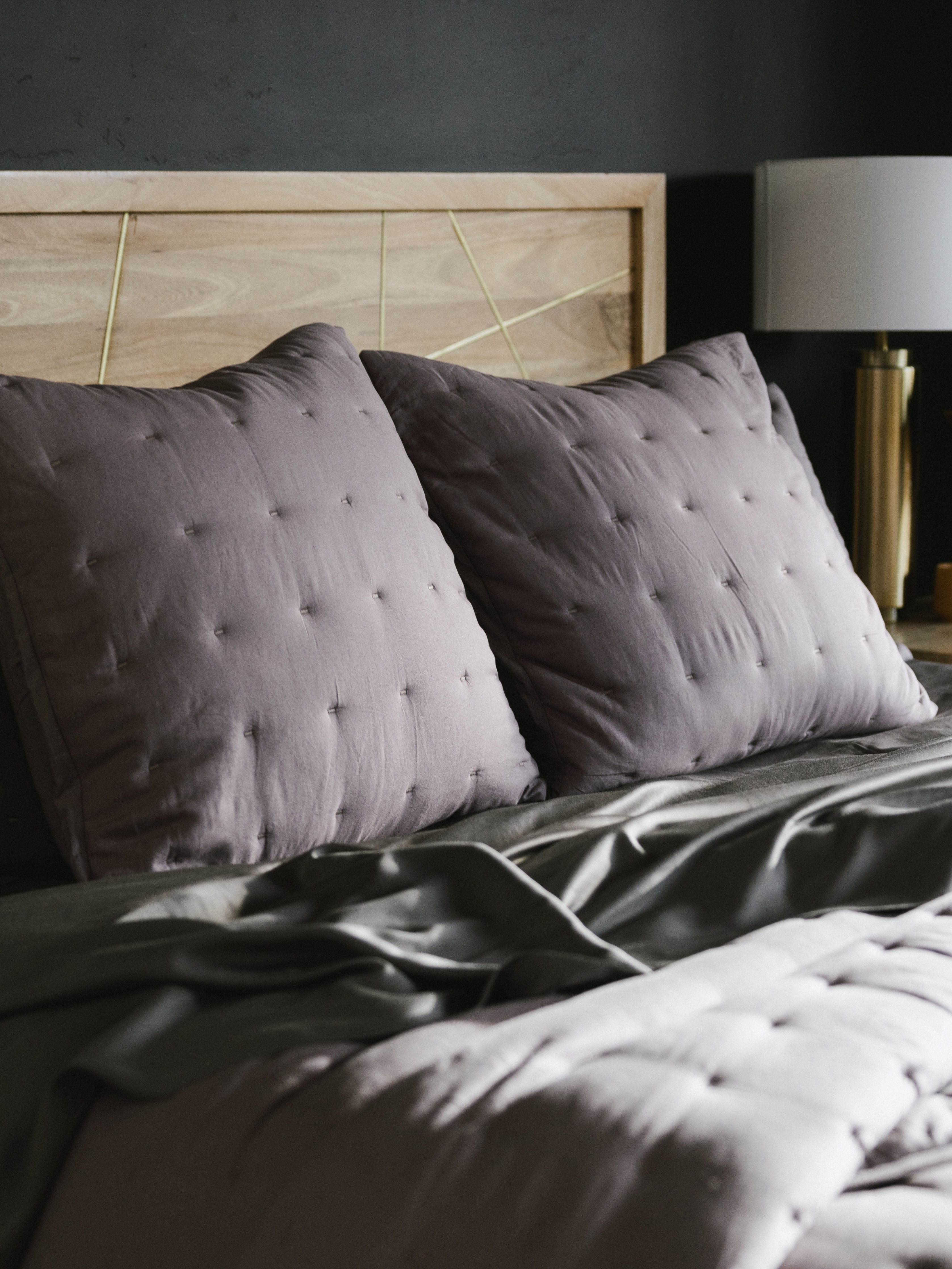 A cozy bed with Cozy Earth's Premium Down Alternative Insert covered in dark gray bedding and adorned with two large, tufted gray pillows rests against a light wood headboard. On the right side, a modern bedside lamp with a gold base adds elegance to the scene. The room's dark wall background creates a striking contrast with the light-colored headboard and pillows.