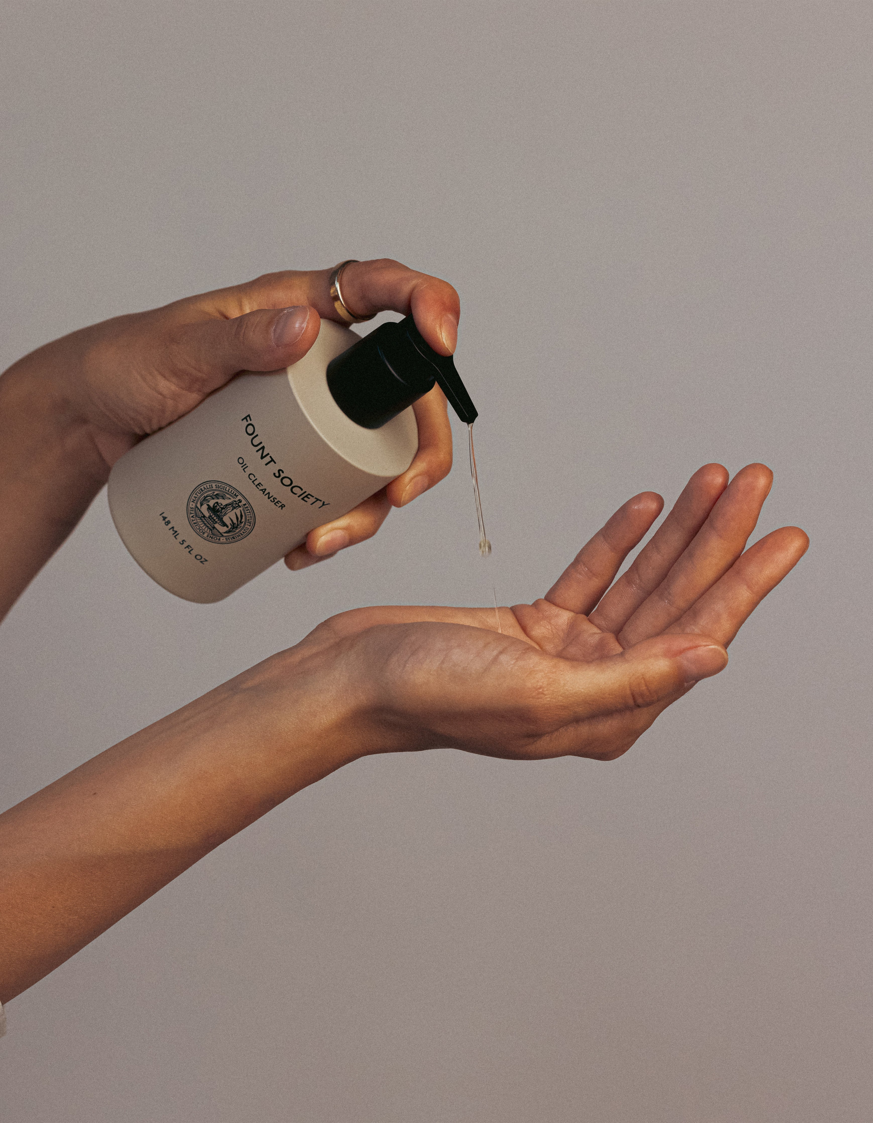 A person is holding a white bottle with a black pump labeled "Cozy Earth Oil Cleanser" and dispensing a drop of liquid onto their other palm, set against a neutral background. The bottle features a round logo, and the hands are positioned gracefully.