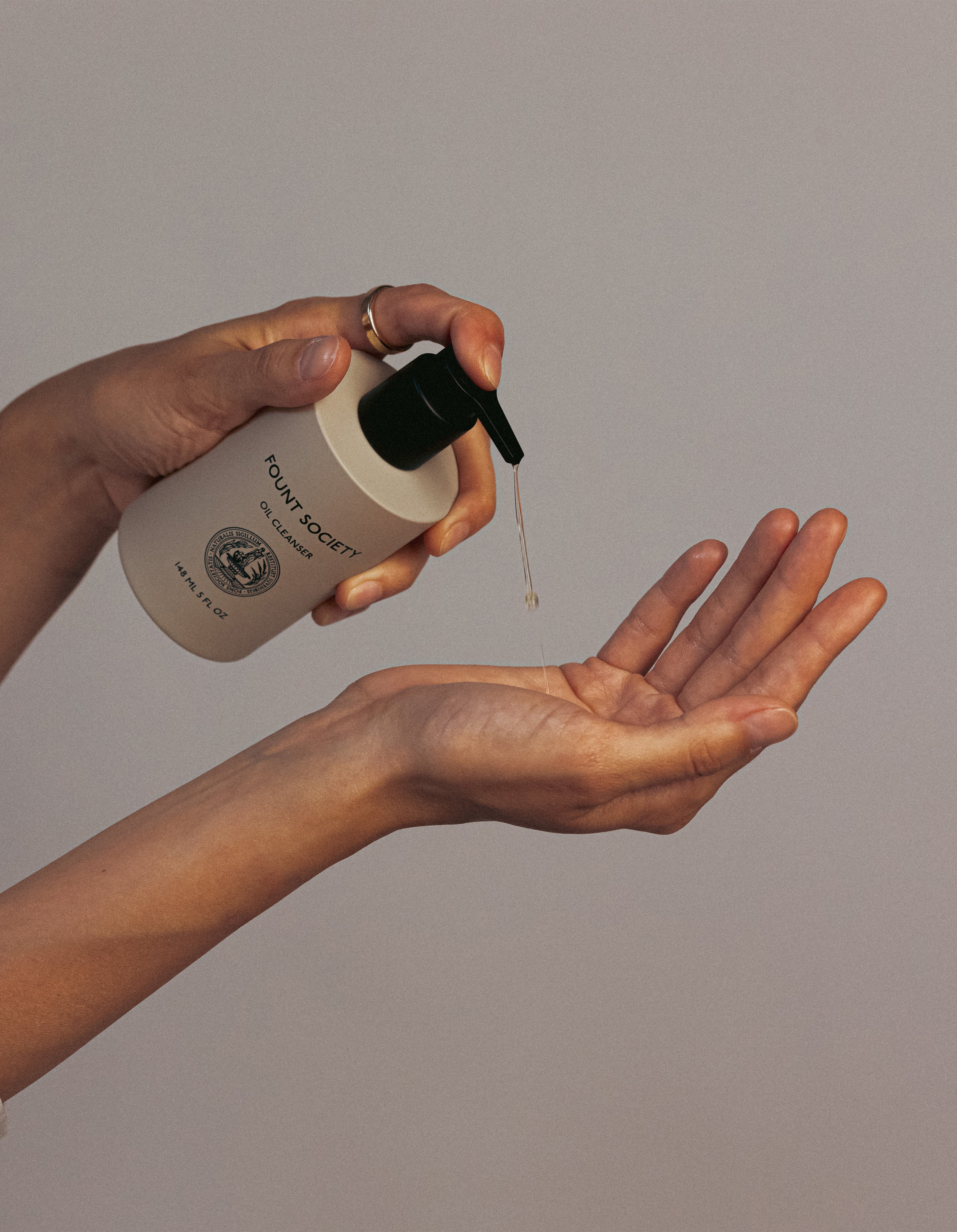 A person dispensing Cozy Earth's Revitalizing Pair from a white bottle labeled "FOUND/SECRET" onto their palm. The bottle has a dark green pump and features an emblem with text. The background is a neutral shade.