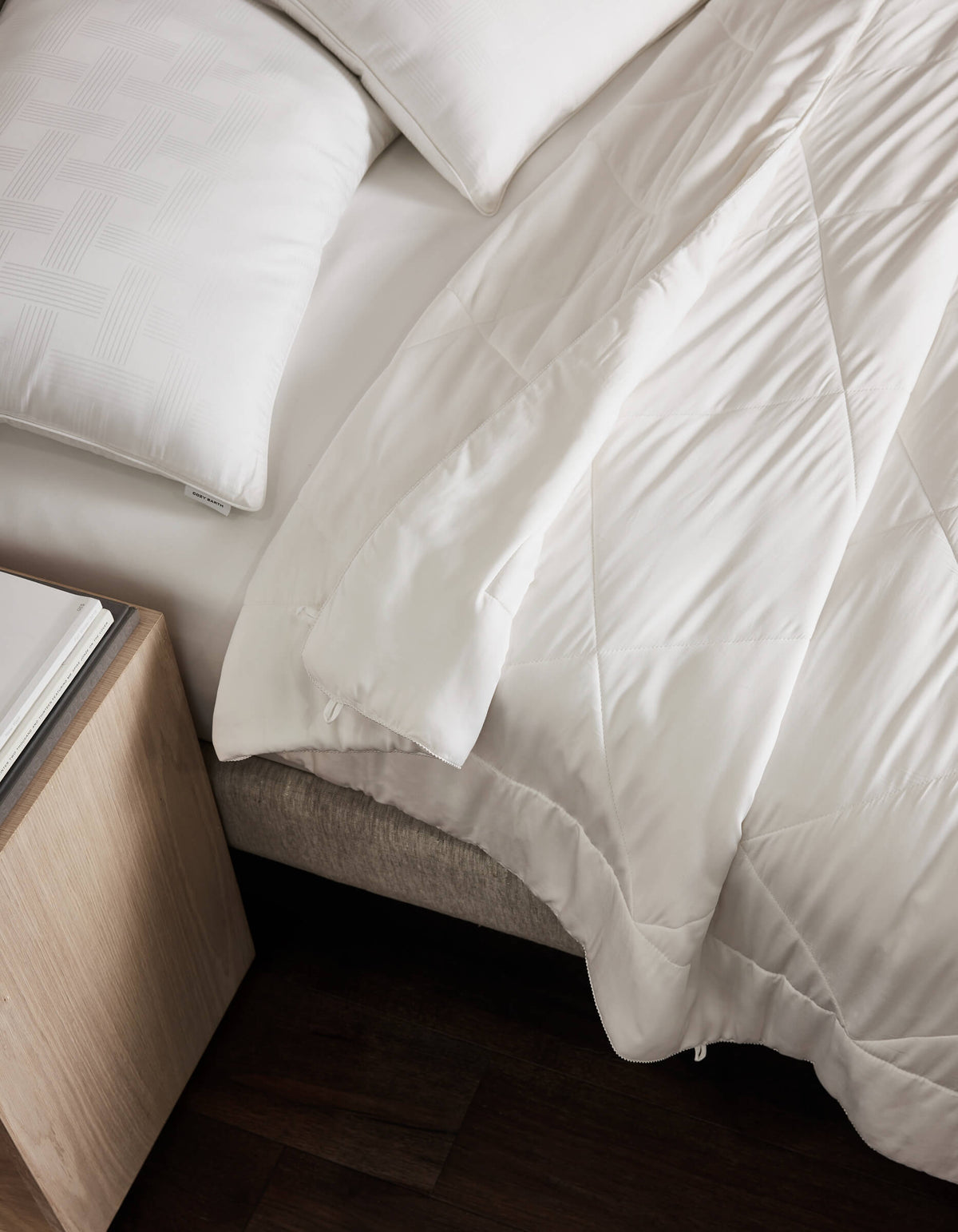A neatly made bed features the Cozy Earth Soft-Wash Cotton Bedding Deluxe Bundle with white linens and pillows, set beside a light wood nightstand on a dark wood floor. The blanket shows a subtle geometric pattern and is slightly folded back.