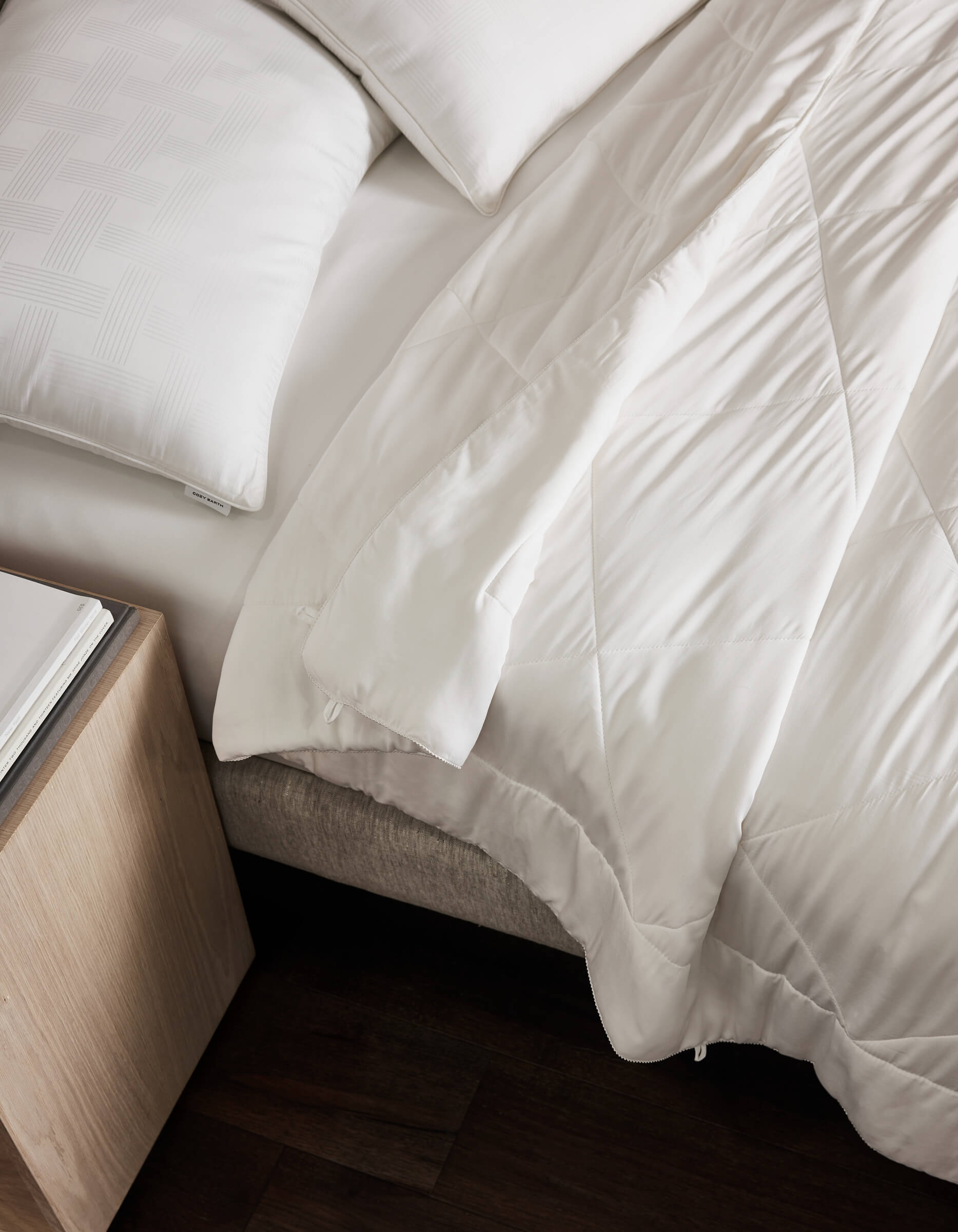 A neatly made bed features the Cozy Earth Soft-Wash Cotton Bedding Deluxe Bundle with white linens and pillows, set beside a light wood nightstand on a dark wood floor. The blanket shows a subtle geometric pattern and is slightly folded back.