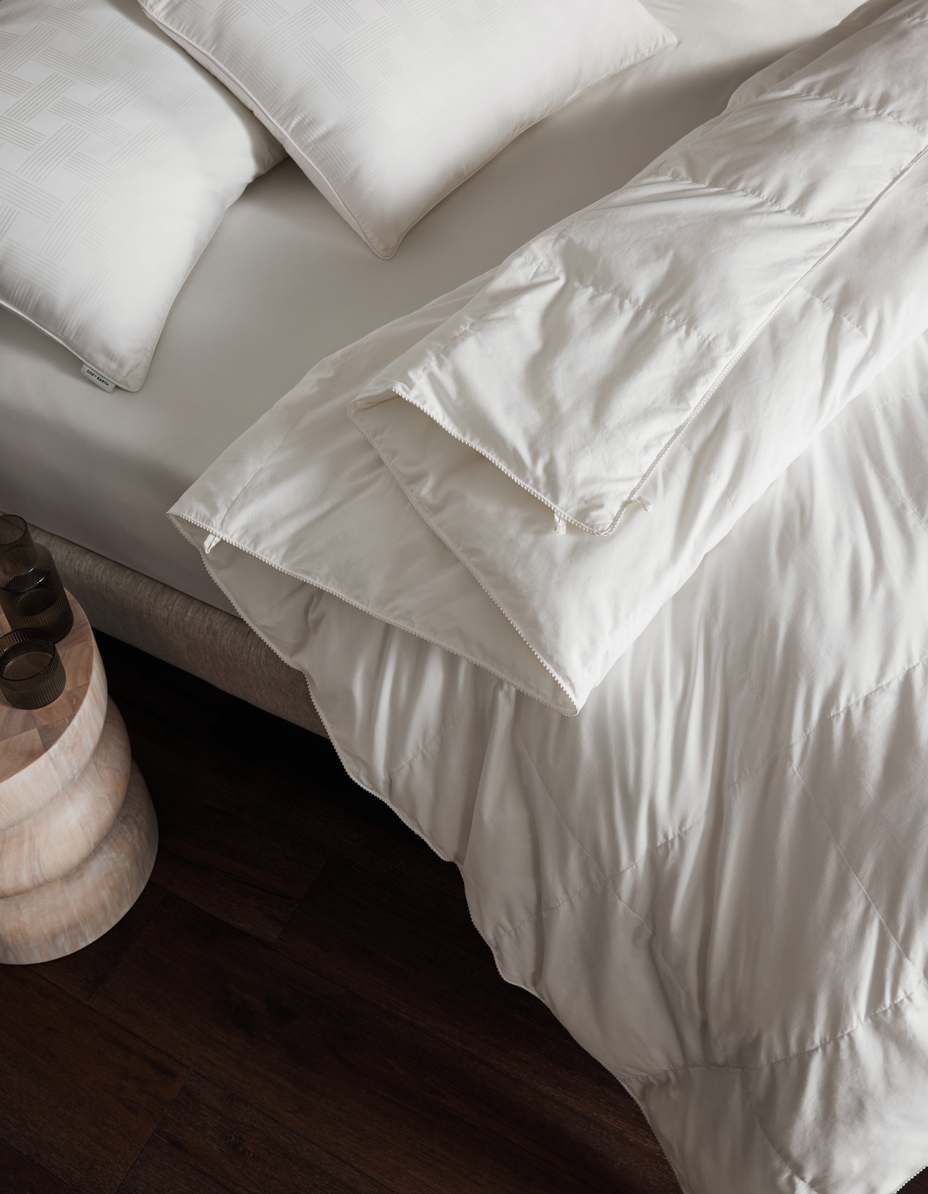 A neatly made bed features Cozy Earth's Down Alternative Comforter and white pillows beside a small wooden nightstand on a dark wood floor.|Filling:Standard