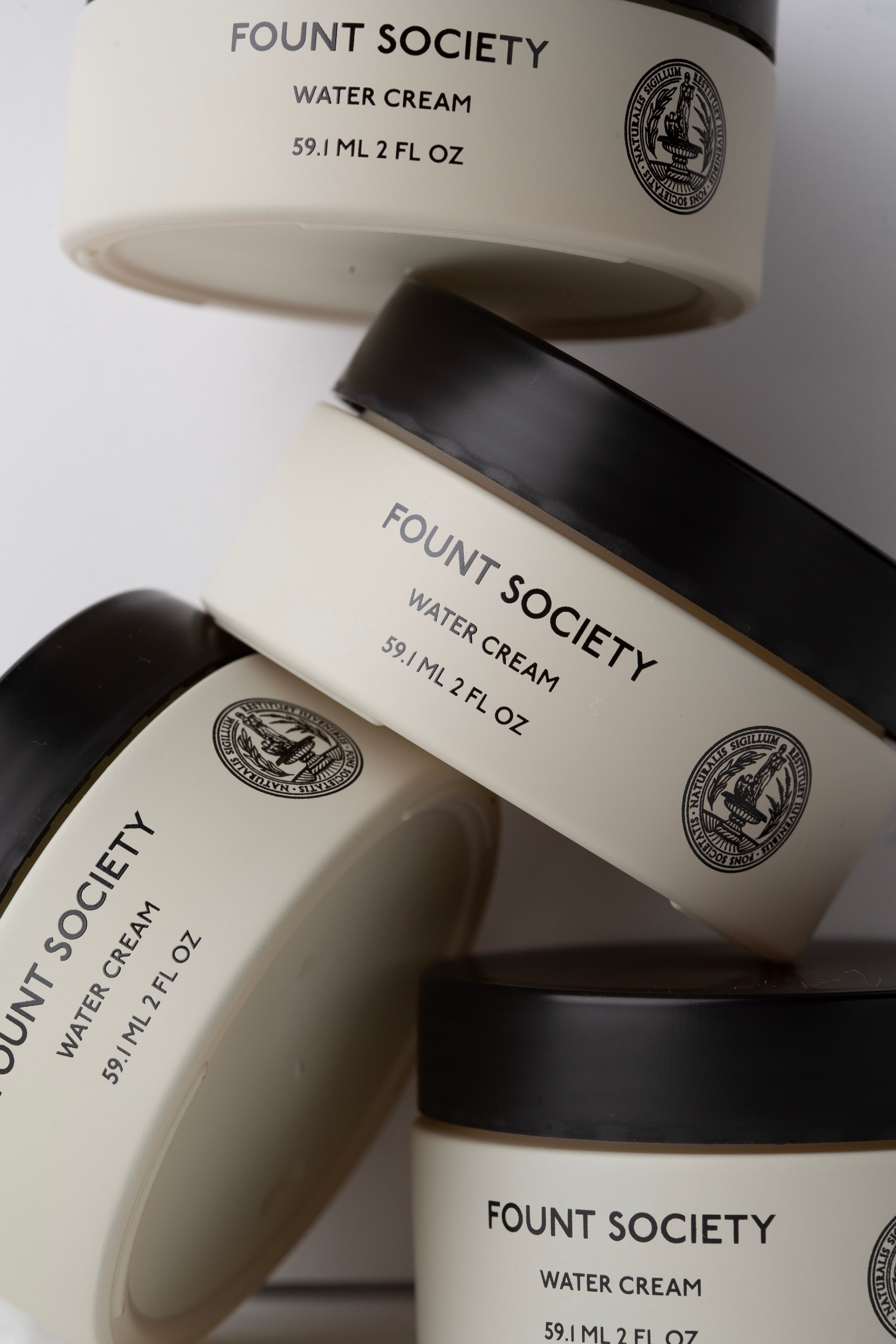 Three jars of Cozy Earth's Water Cream Moisturizer are displayed. The jars are beige with black lids and feature black text and a circular logo. Each jar is labeled with "59.1 ML/2 FL OZ." The arrangement is slightly tilted, showcasing different angles of the products. |Size:Full Size