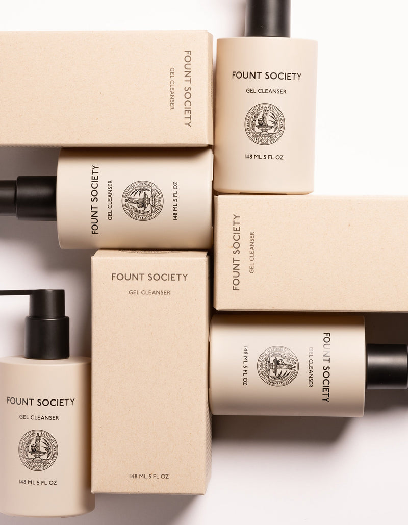 A flat lay of Cozy Earth Gel Cleanser bottles and their beige packaging boxes arranged in a neat, geometric pattern. The cylindrical bottles have black pumps and white labels with black text, stating the product details and volume (148ml/5 fl oz).