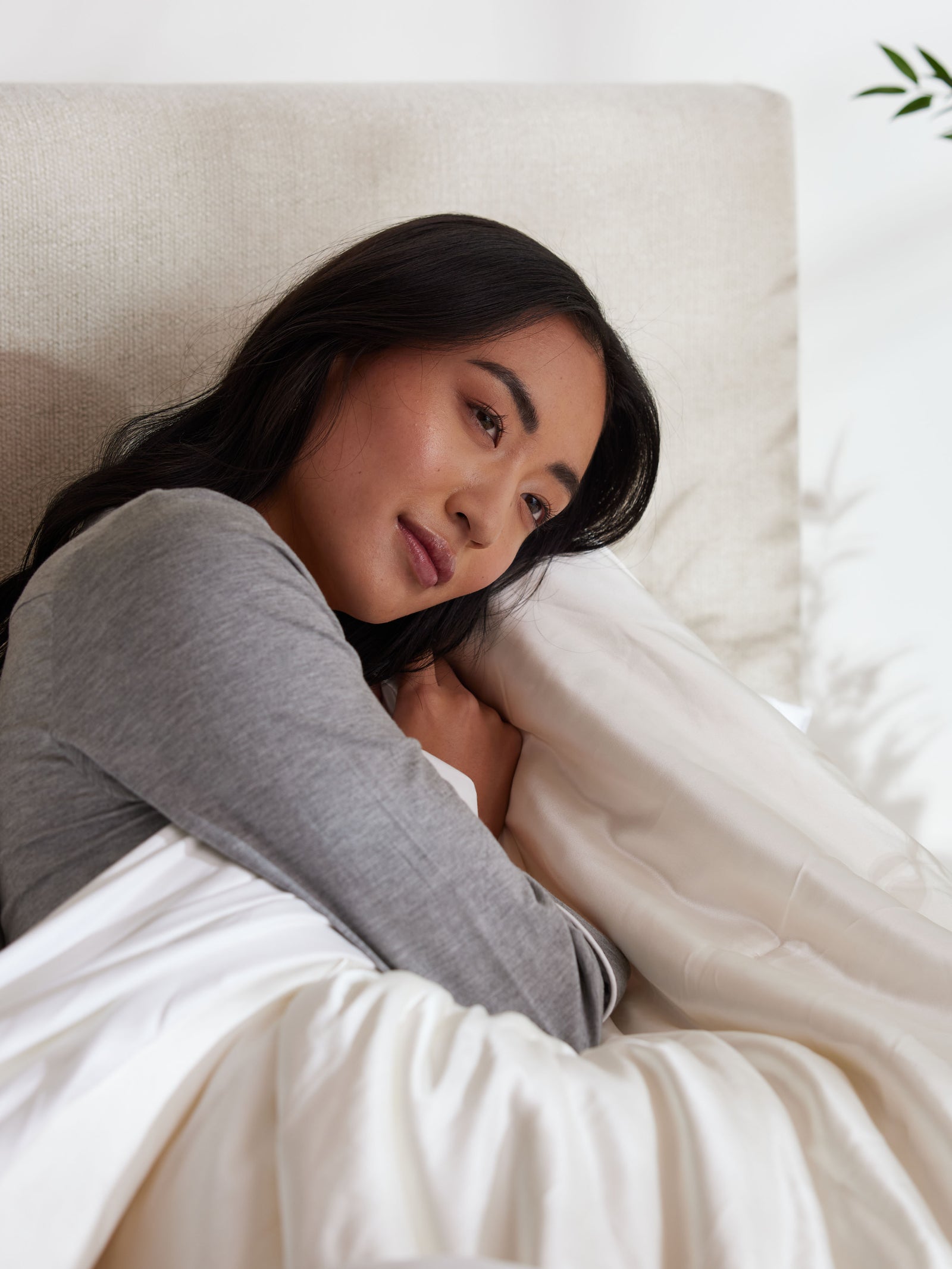 A person with long, dark hair lies in bed, enveloped in a Silk Comforter by Cozy Earth. They are wearing a gray long-sleeve top and appear relaxed, gazing thoughtfully into the distance.