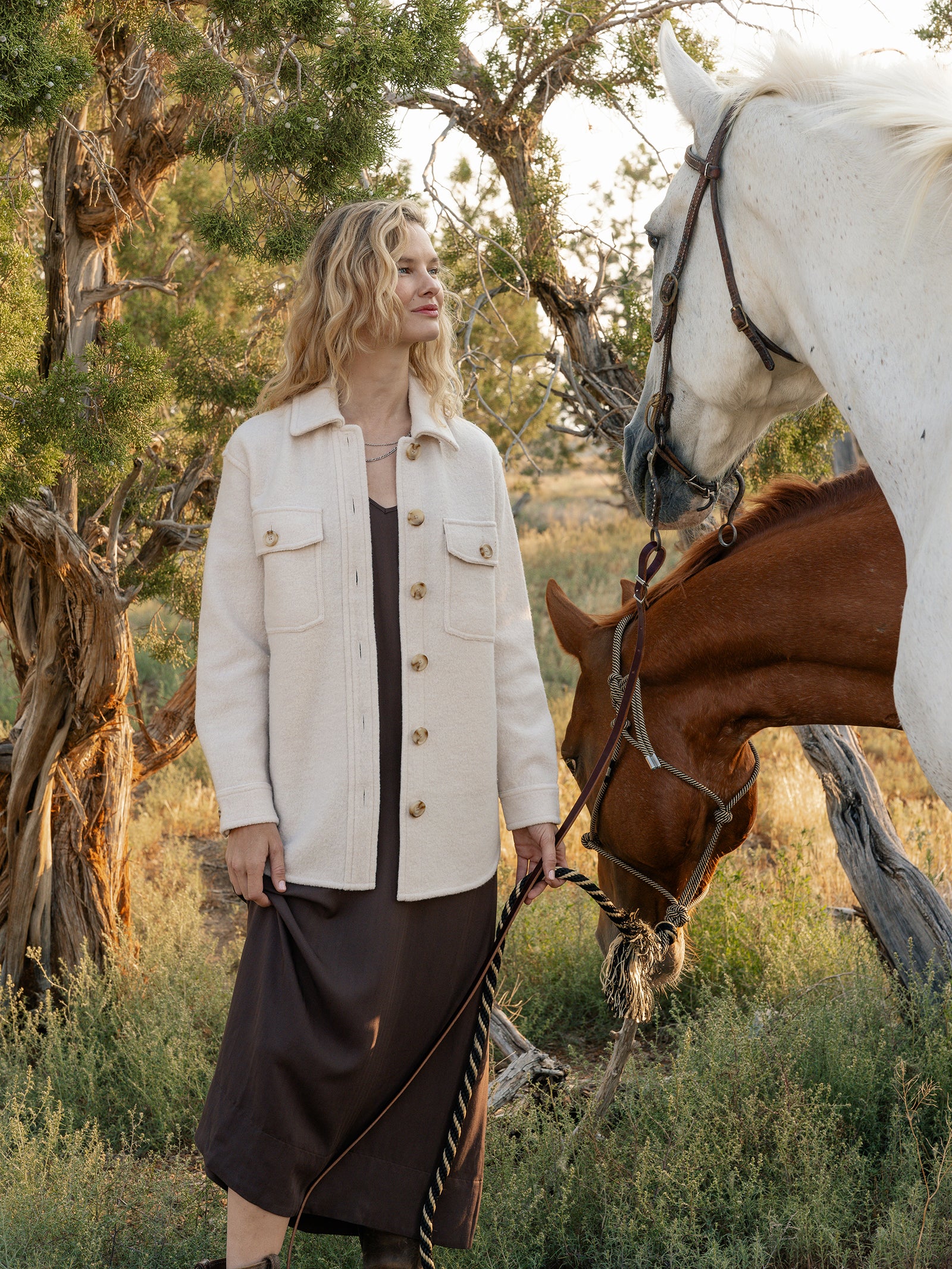 A person with wavy blonde hair stands outdoors wearing a Cozy Earth's Women's Boucle Shacket in beige over a brown dress, holding the reins of two horses. The background features trees and grass, suggesting a natural setting.  