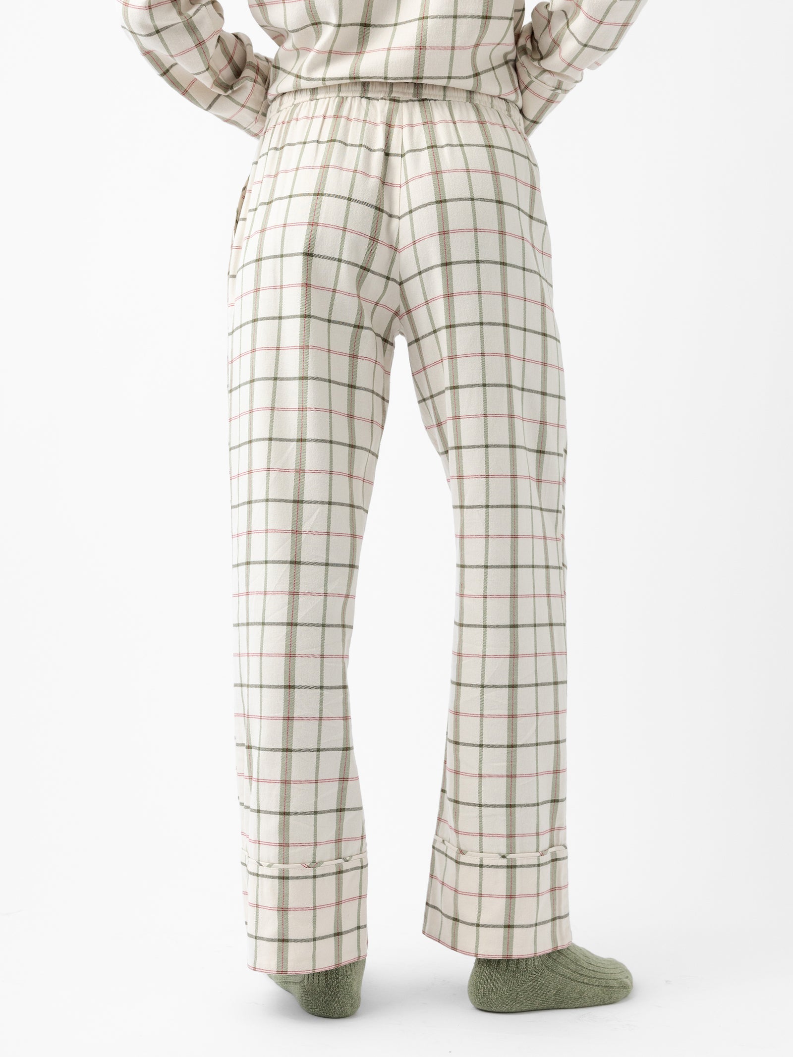 A person wearing Cozy Earth's Women's Farmhouse Long Sleeve Pajama Set stands with their back to the camera. The pajamas feature a predominantly white fabric with a grid pattern of green and pinkish-red lines. The bottom of the pajama pants is slightly gathered, and the person is also wearing green socks. 