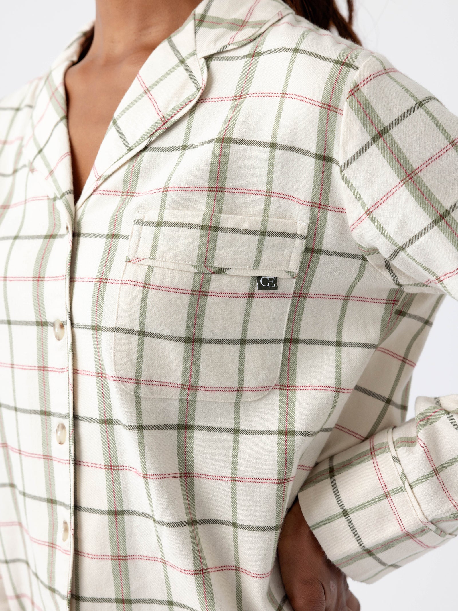 A person wearing a cream-colored plaid shirt from the Cozy Earth Women's Farmhouse Long Sleeve Pajama Set, featuring green and pink lines, stands with one hand on their hip. The shirt includes a button-up front and a chest pocket with a small tag. The background is plain white. 