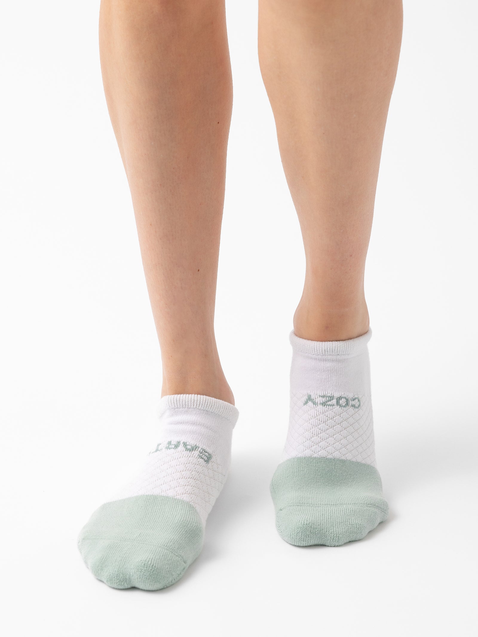 A person wearing a pair of white Essential Ankle Socks by Cozy Earth, featuring light green toes and the text "COZY" visible on one sock, is standing against a plain white background. 