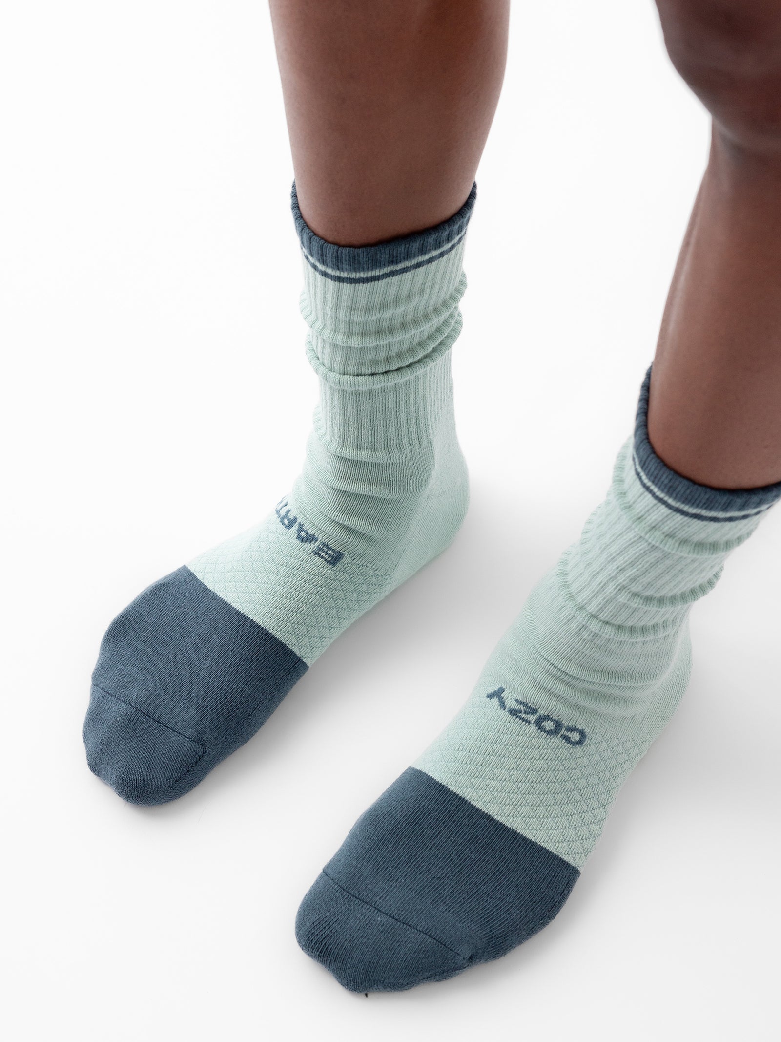 Close-up image of a person's lower legs and feet wearing light green Essential Calf Socks by Cozy Earth, featuring dark green accents on the toes, heels, and cuffs. The socks have "EARTH" and "COZY" printed near the toes. The person is standing on a white background. 