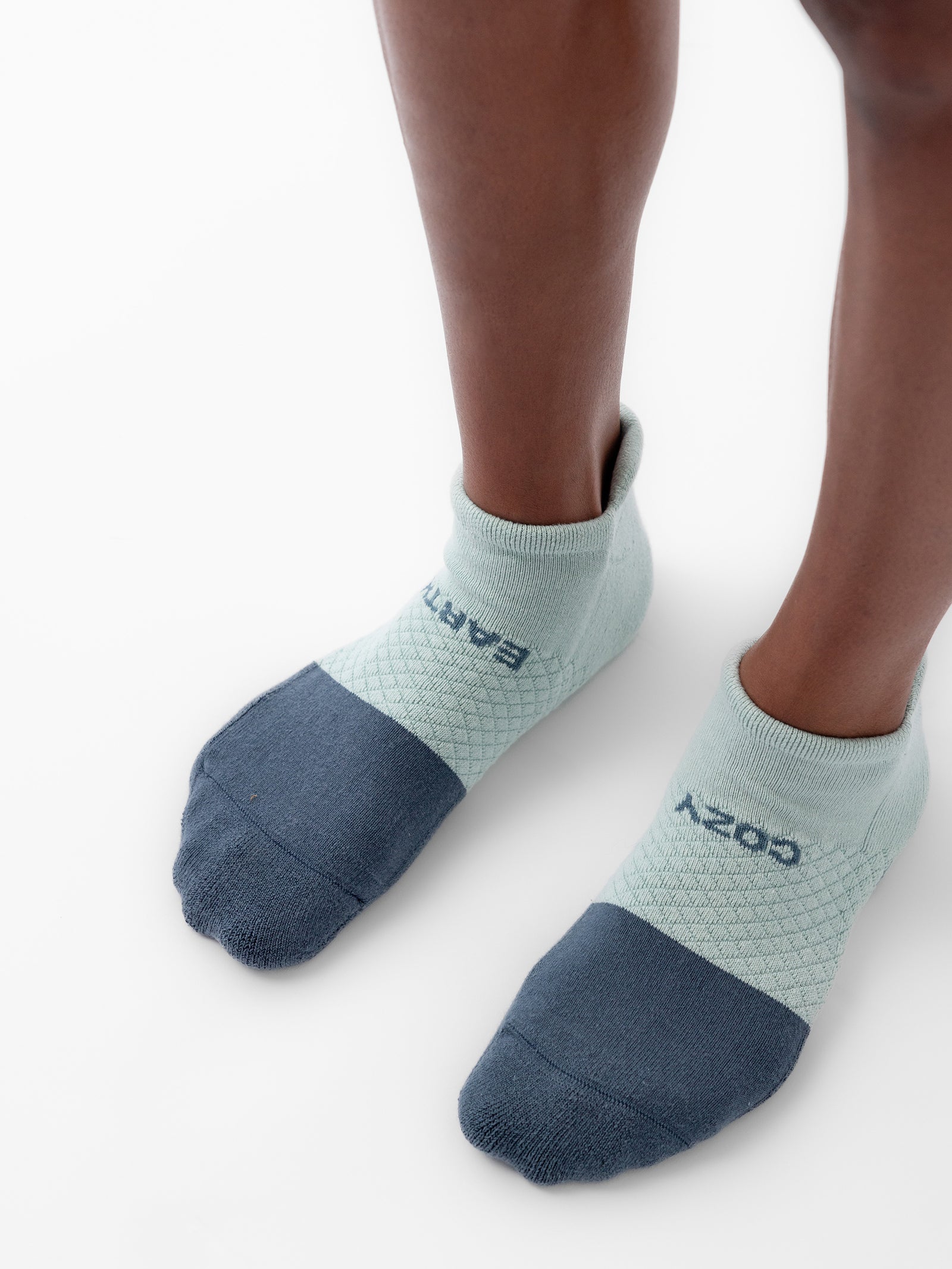 A person wearing a pair of color-blocked socks with light green tops and dark gray toes from the Cozy Earth's Essential Ankle Sock 4-Pack. The socks have the words "COZY" and "ARTFUL" written on them in dark letters, while the person stands on a white surface. 