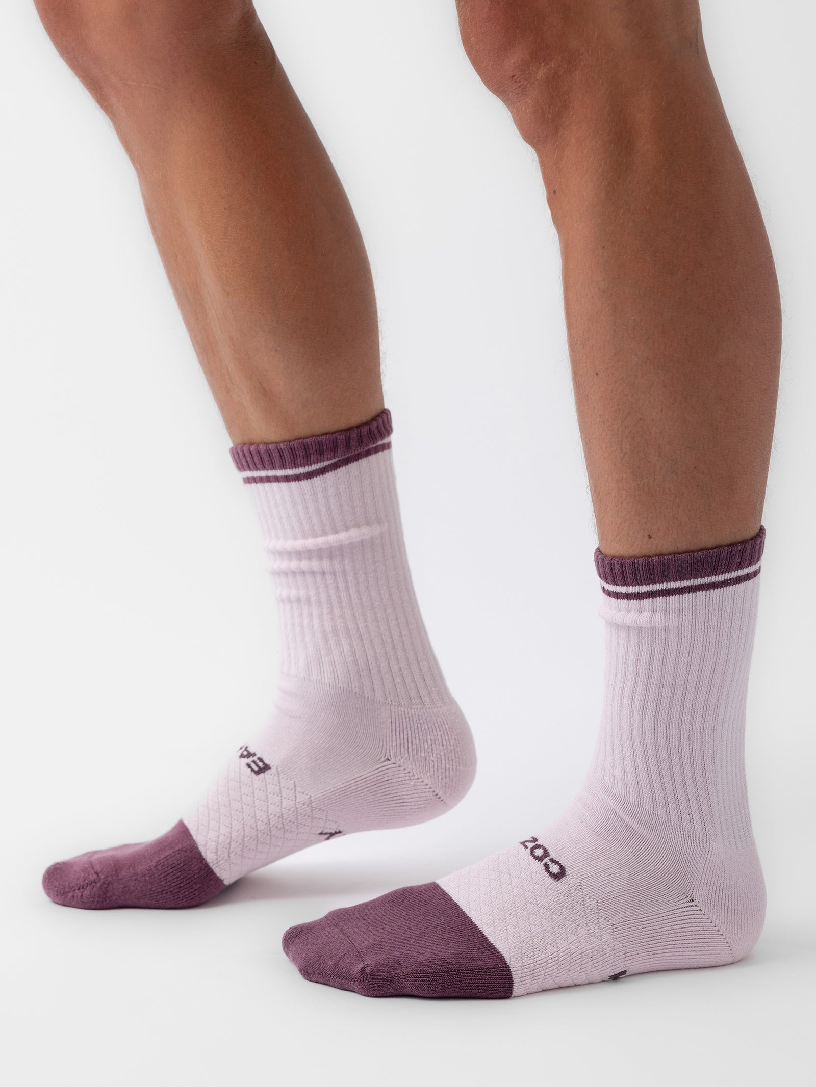 A person is wearing white crew socks from the Essential Calf Sock 4-Pack by Cozy Earth, which feature purple detailing on the toes, heels, and cuffs. The socks have a ribbed texture around the ankles, and the letters "CCD" are visible on the sides of the socks. The background is white. 