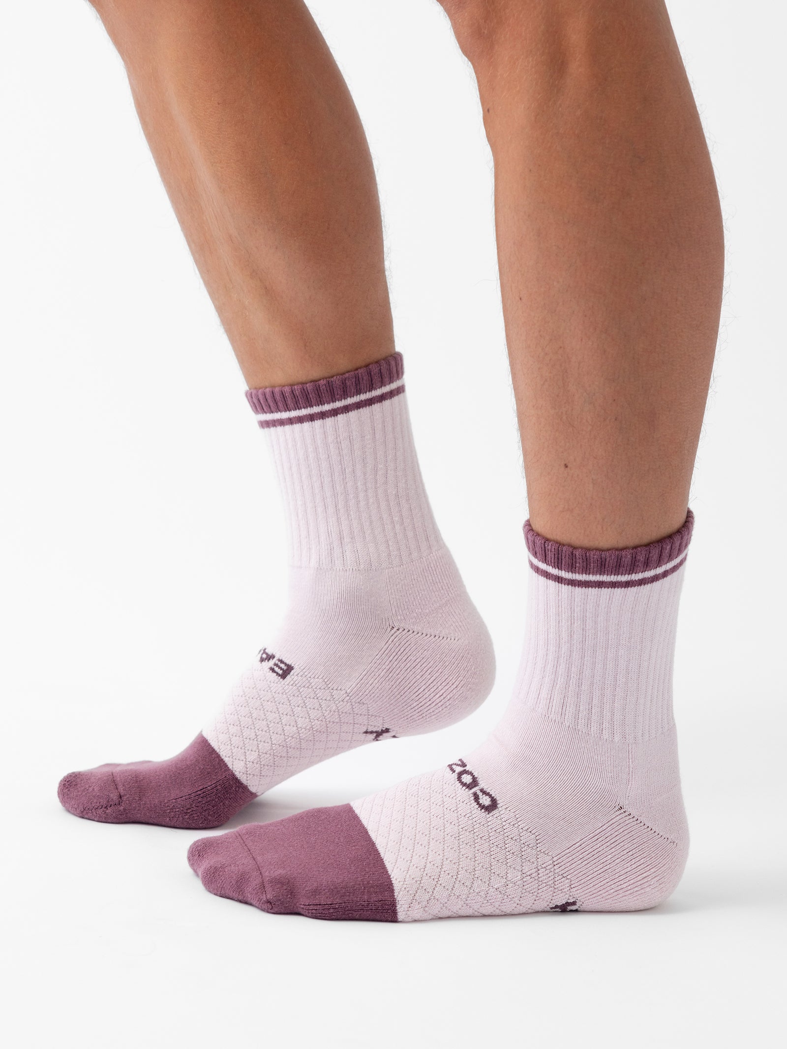 A pair of legs wearing light pink Essential Quarter Socks from Cozy Earth's 4-Pack, adorned with dark purple toes, heels, and upper rims. The socks boast a textured pattern and the word "Balega" alongside another partially visible word on the sides. The background is plain white. 