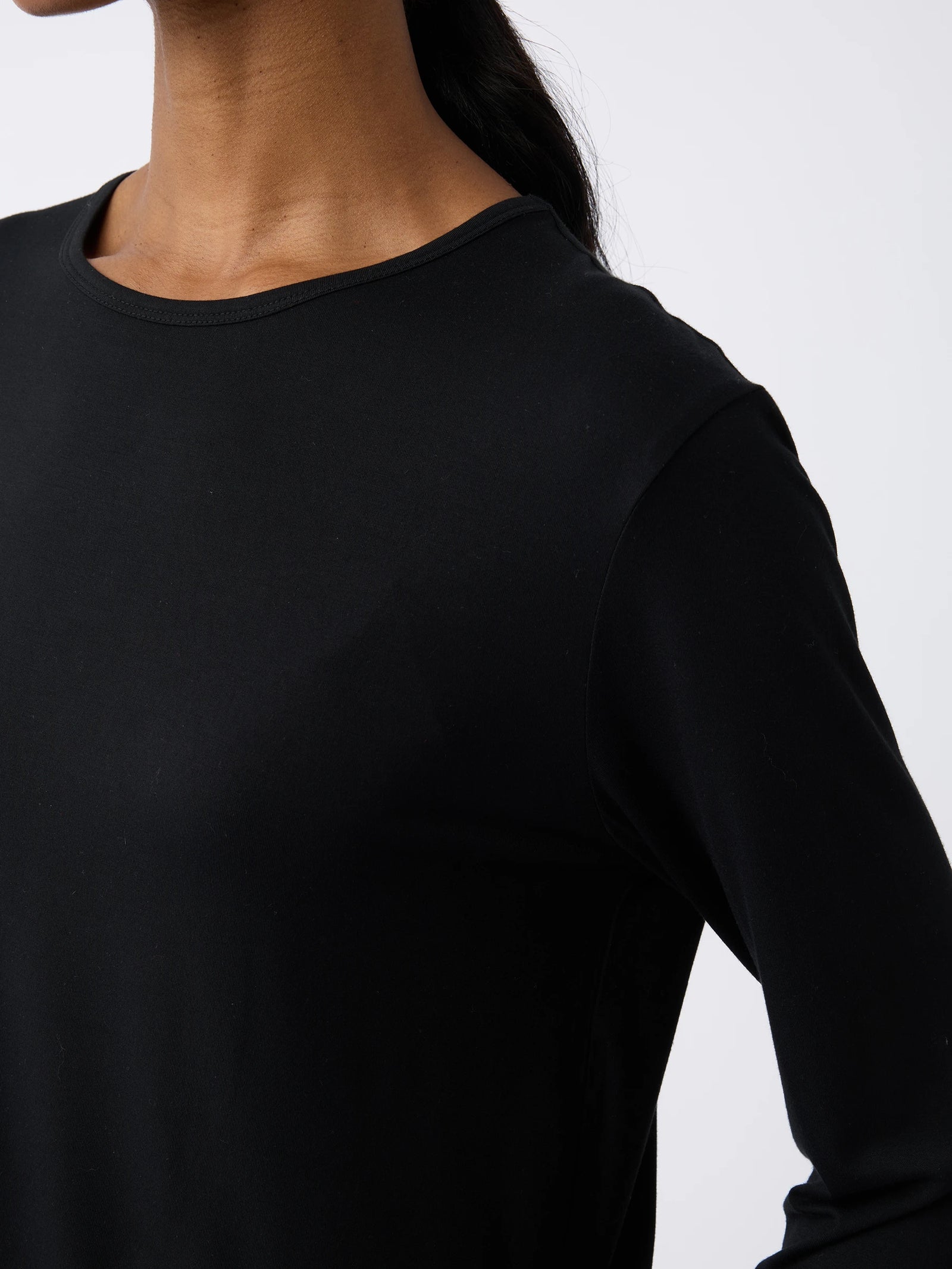 A model in a black Women's Stretch Knit Bamboo Long Sleeve Lounge Tee by Cozy Earth is shown from neck to waist against a white background, her head slightly turned away from the camera. 