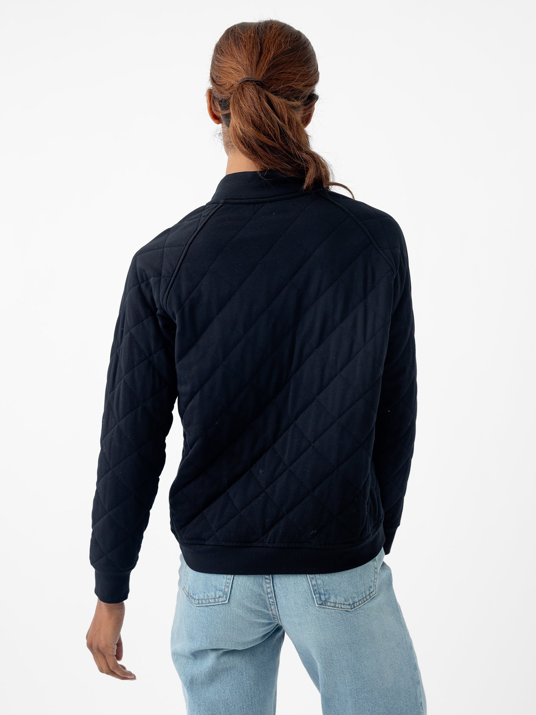 A person with long hair tied in a ponytail is seen from behind, wearing a black Women's Ultra-Soft Bamboo Quilted Snap Pullover by Cozy Earth and light blue jeans. The background is plain white. 