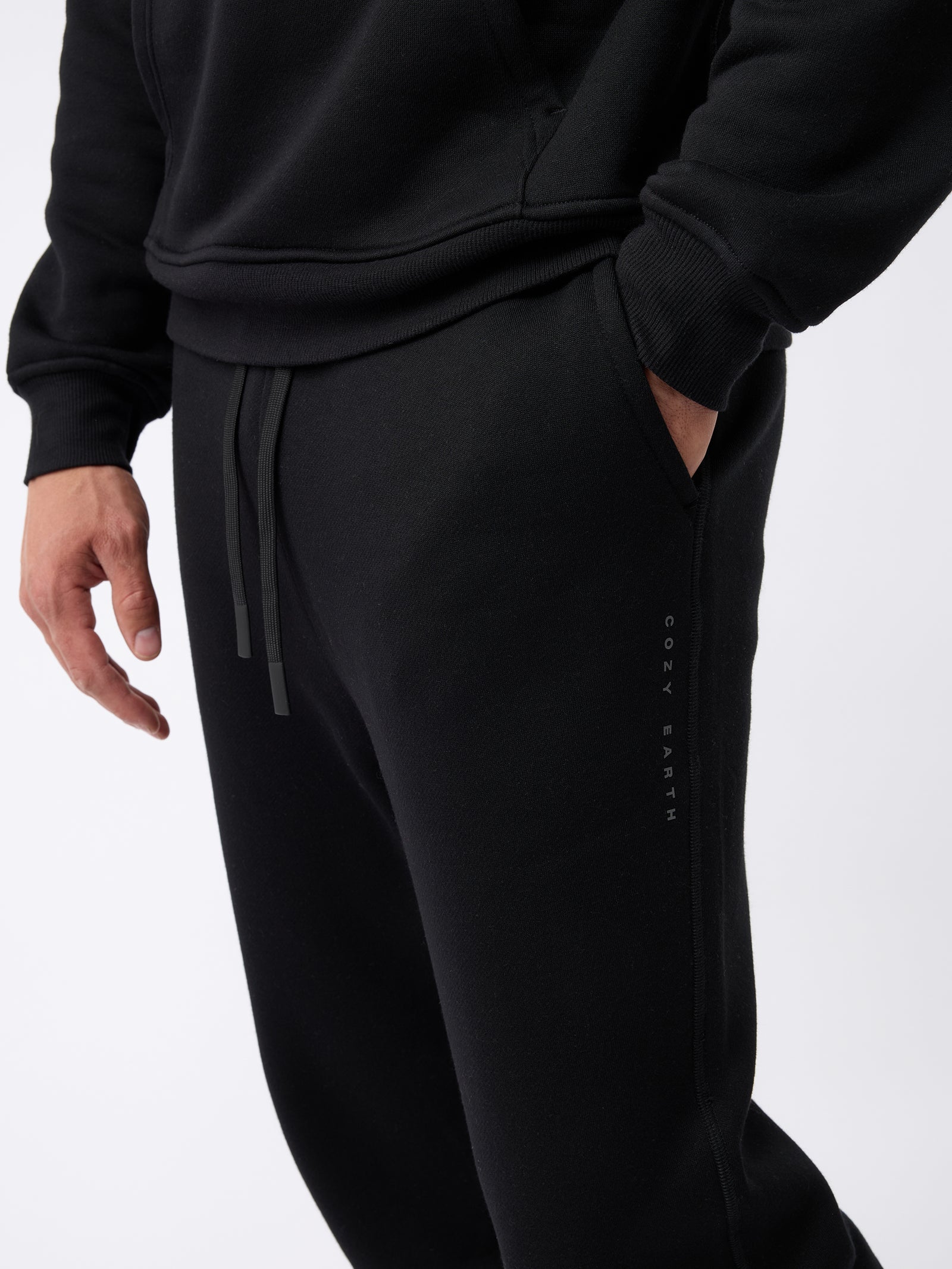 A person is wearing a black hoodie and the Men's CityScape Jogger in black, with their left hand in the jogger's pocket. The brand name "Cozy Earth" is visible on the side of the joggers against a plain white background. 