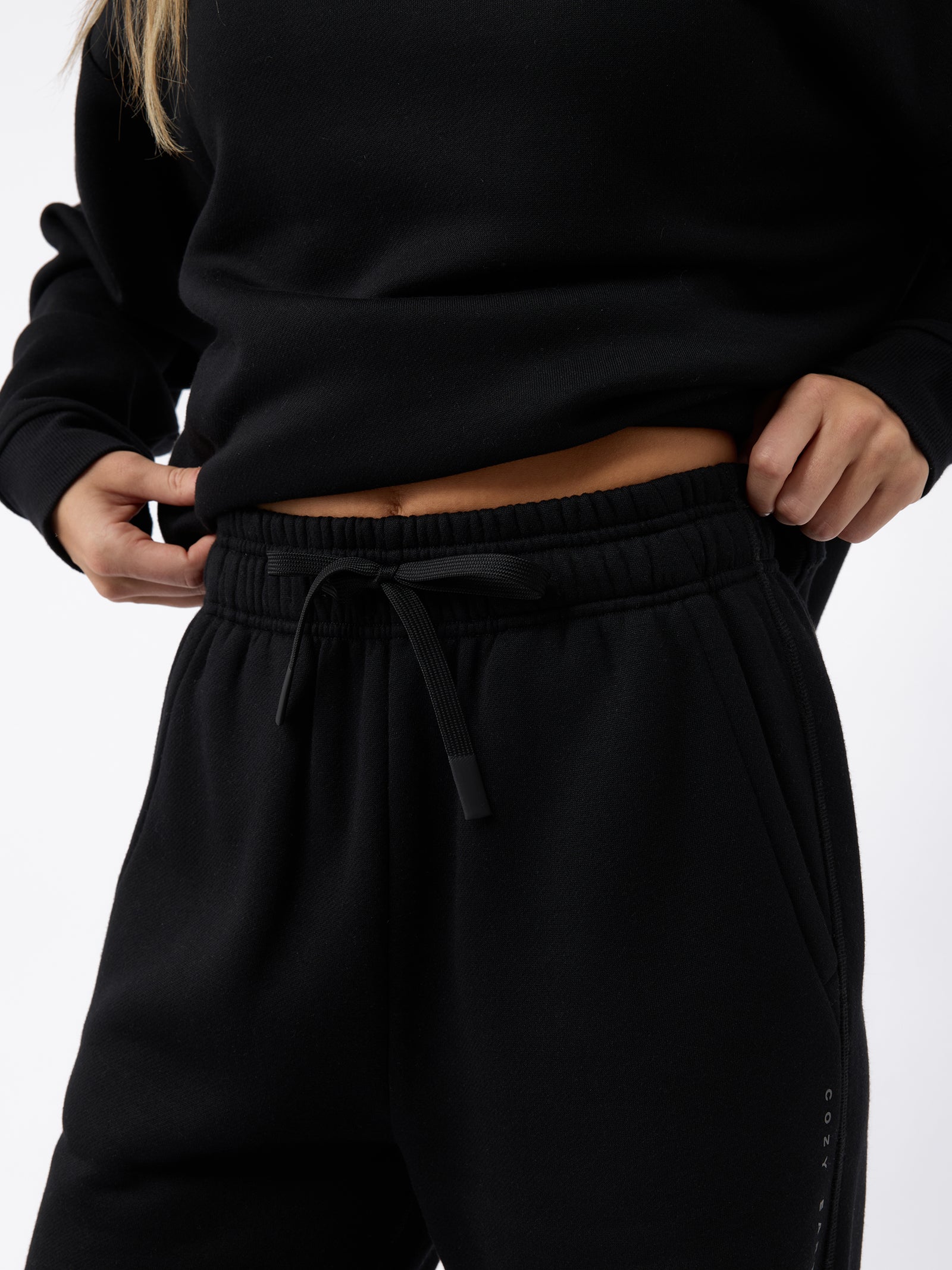 A person wearing a black Women's CityScape Wide Leg Pant from Cozy Earth adjusts the waistband. The photo focuses on their midsection, highlighting the drawstring and fabric texture against a plain white background. 