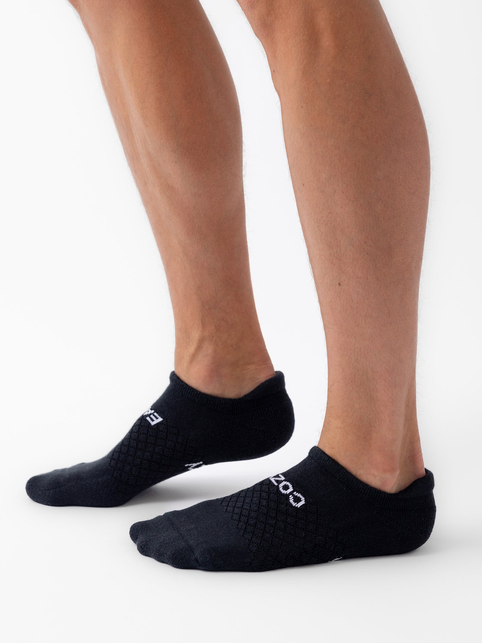 A person wearing black socks from the Essential Ankle Sock 2-Pack by Cozy Earth stands on a white background. The socks feature a textured pattern and the word "COZY" on the sides. The person’s lower legs are visible, and they are standing with one foot slightly in front of the other. 