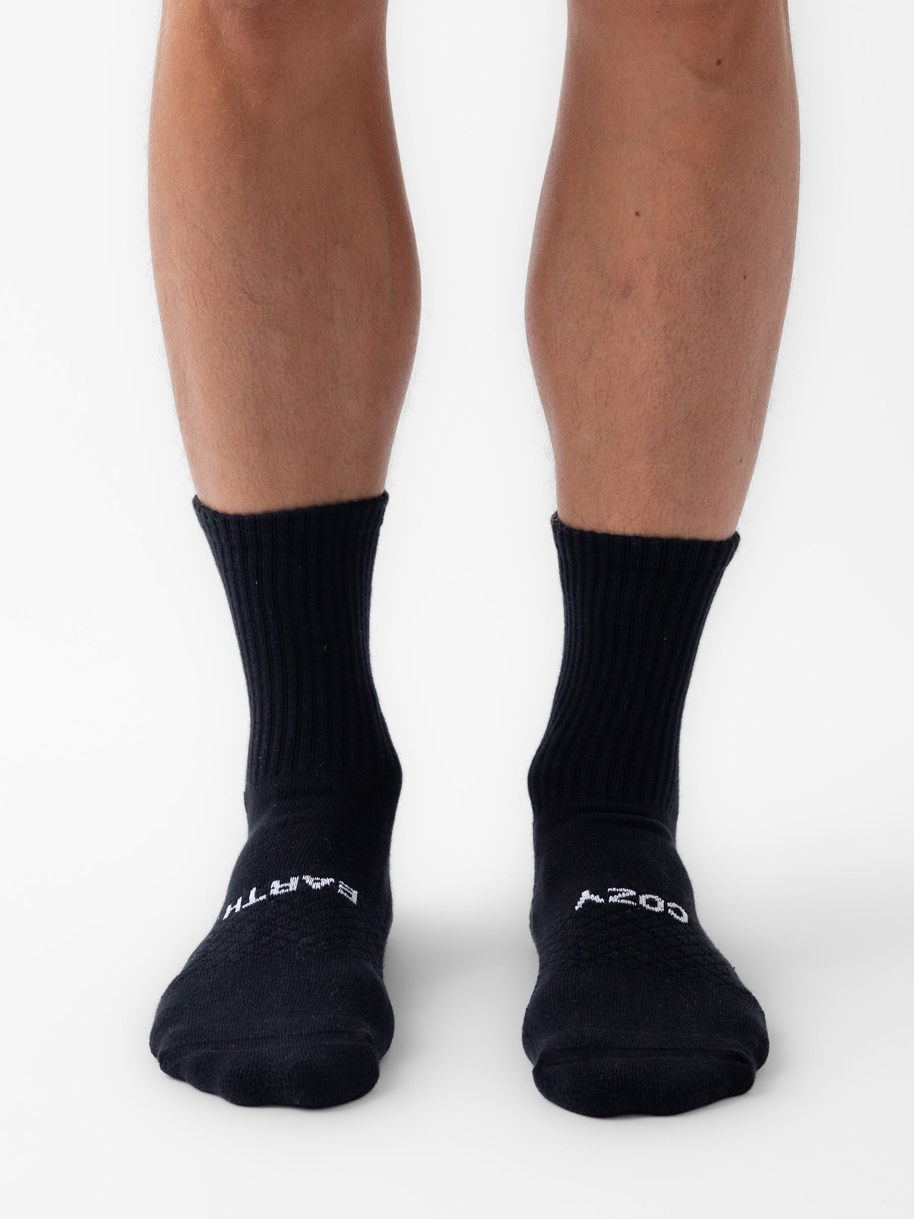 A person stands on a white surface wearing Cozy Earth Essential Calf Sock 8-Pack black crew socks, showing only their legs from the knees down. The socks feature white text near the toes. 
