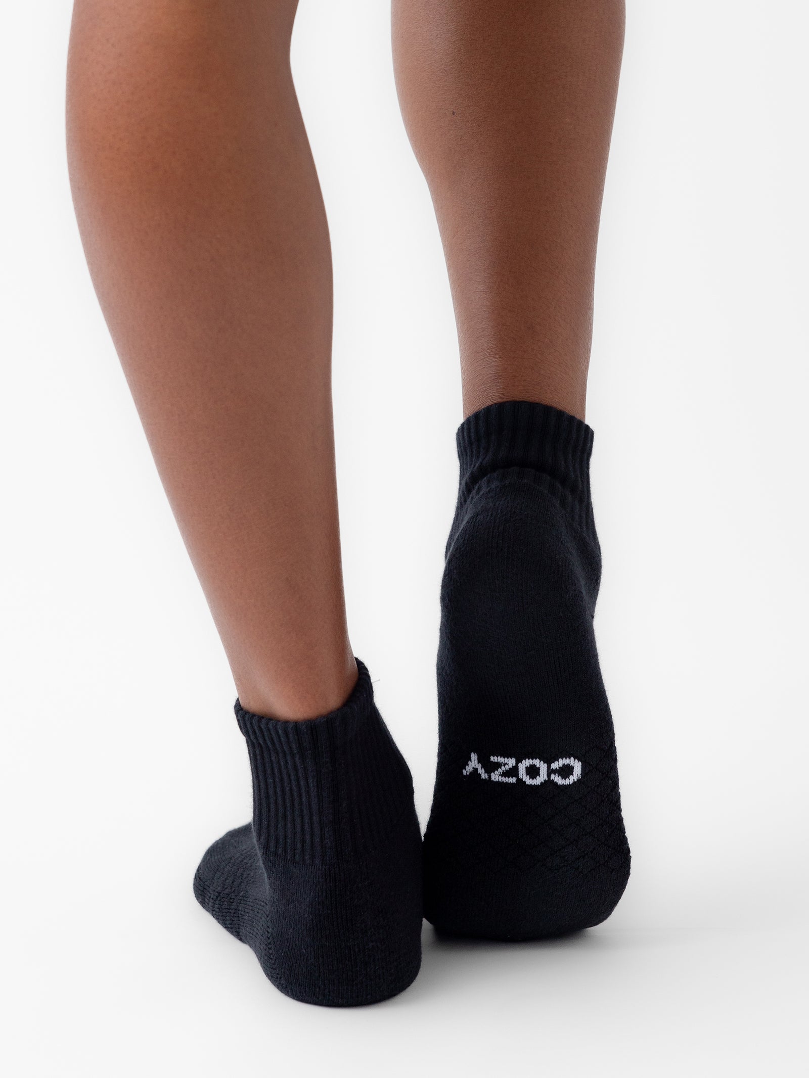 A person stands on their tiptoes, revealing the back of their legs and feet adorned with black Essential Quarter Socks from Cozy Earth. The left sock features the word "COZY" printed in white letters on the sole, set against a plain white background. 