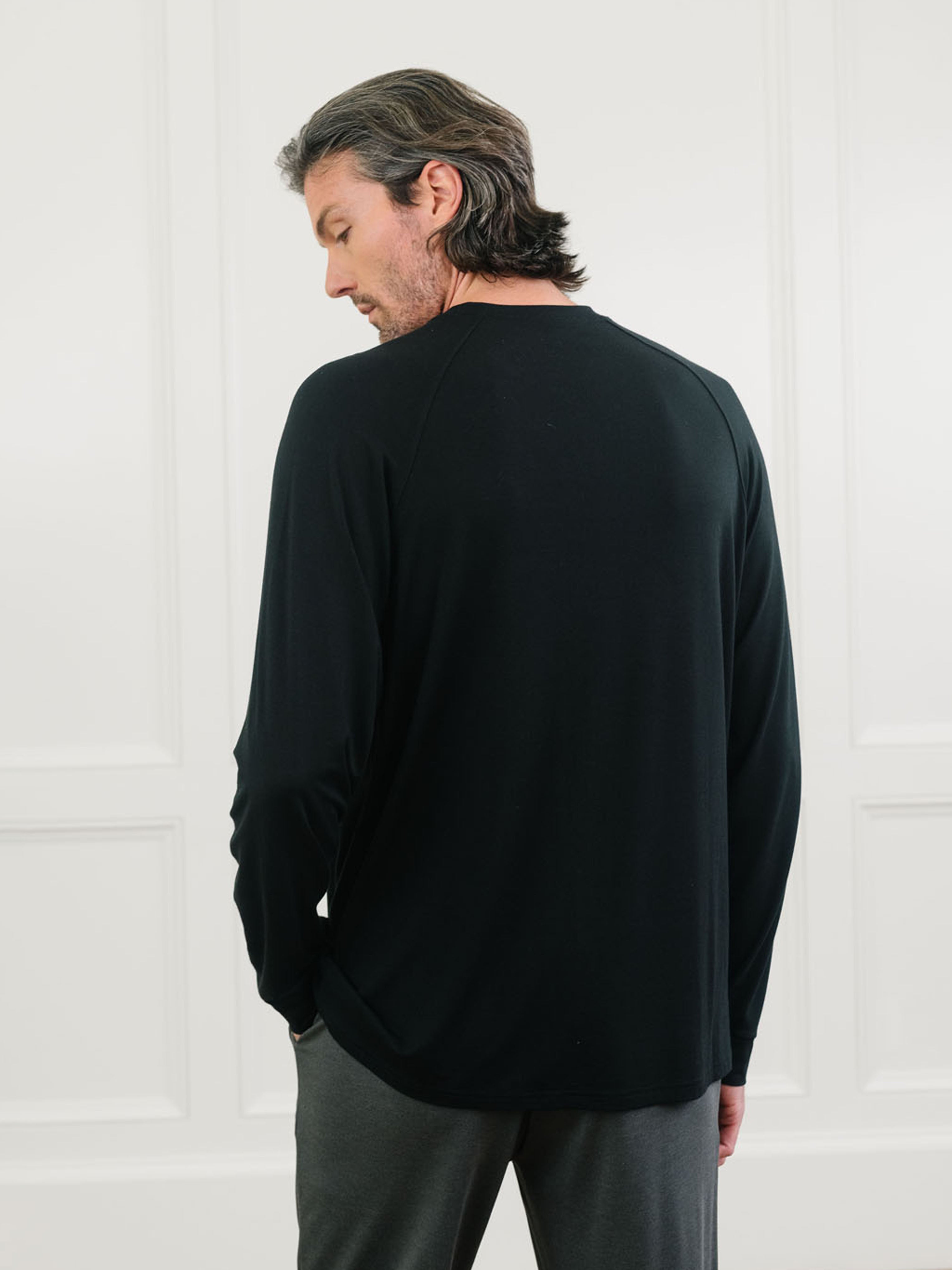 A man with medium-length dark hair, dressed in a Cozy Earth Men's Stretch-Knit Long Sleeve Bamboo Pajama Set, stands facing away from the camera in a well-lit room with white paneled walls.  |Color:Black