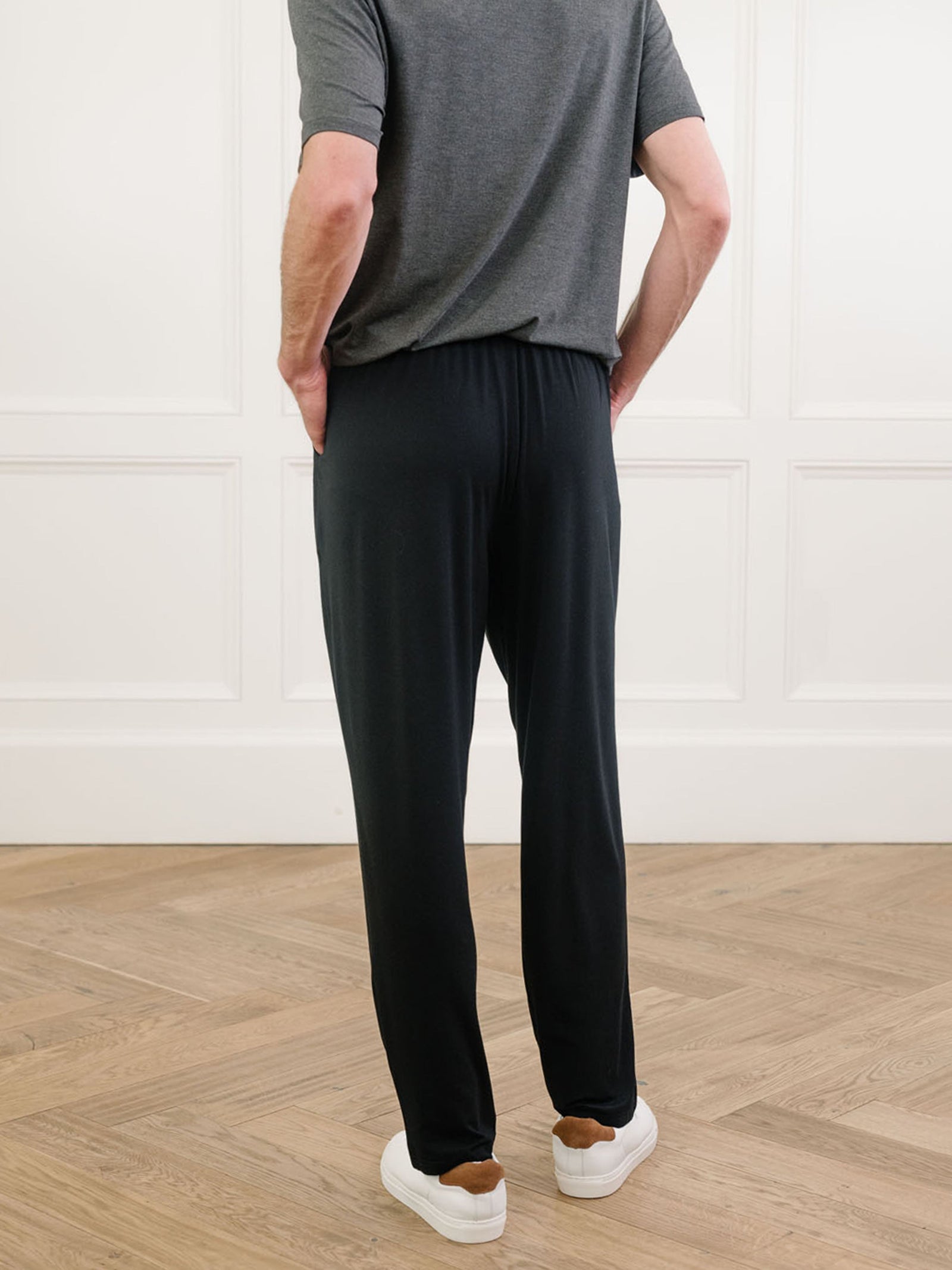 A person wearing the Men's Stretch-Knit Long Sleeve Bamboo Pajama Set in dark grey from Cozy Earth and black pants stands with hands in pockets, showing their back. They are inside a room with light-colored paneled walls and a wooden floor, wearing white shoes.  