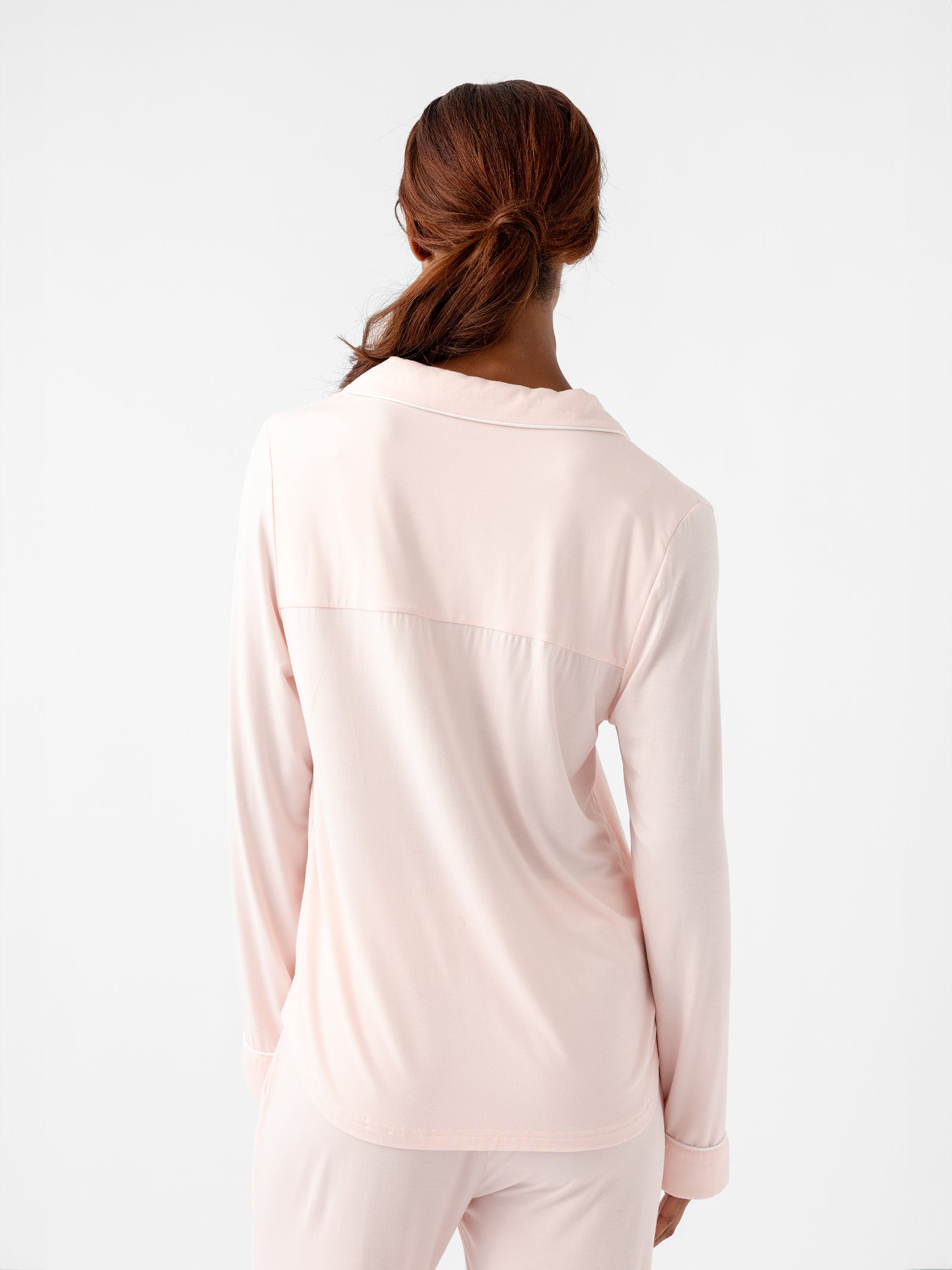 A person with long, brown hair tied in a ponytail is standing with their back to the camera. They are wearing the Women's Long Sleeve Bamboo Pajama Top in Stretch-Knit TALL by HIDE. The background is plain white. 