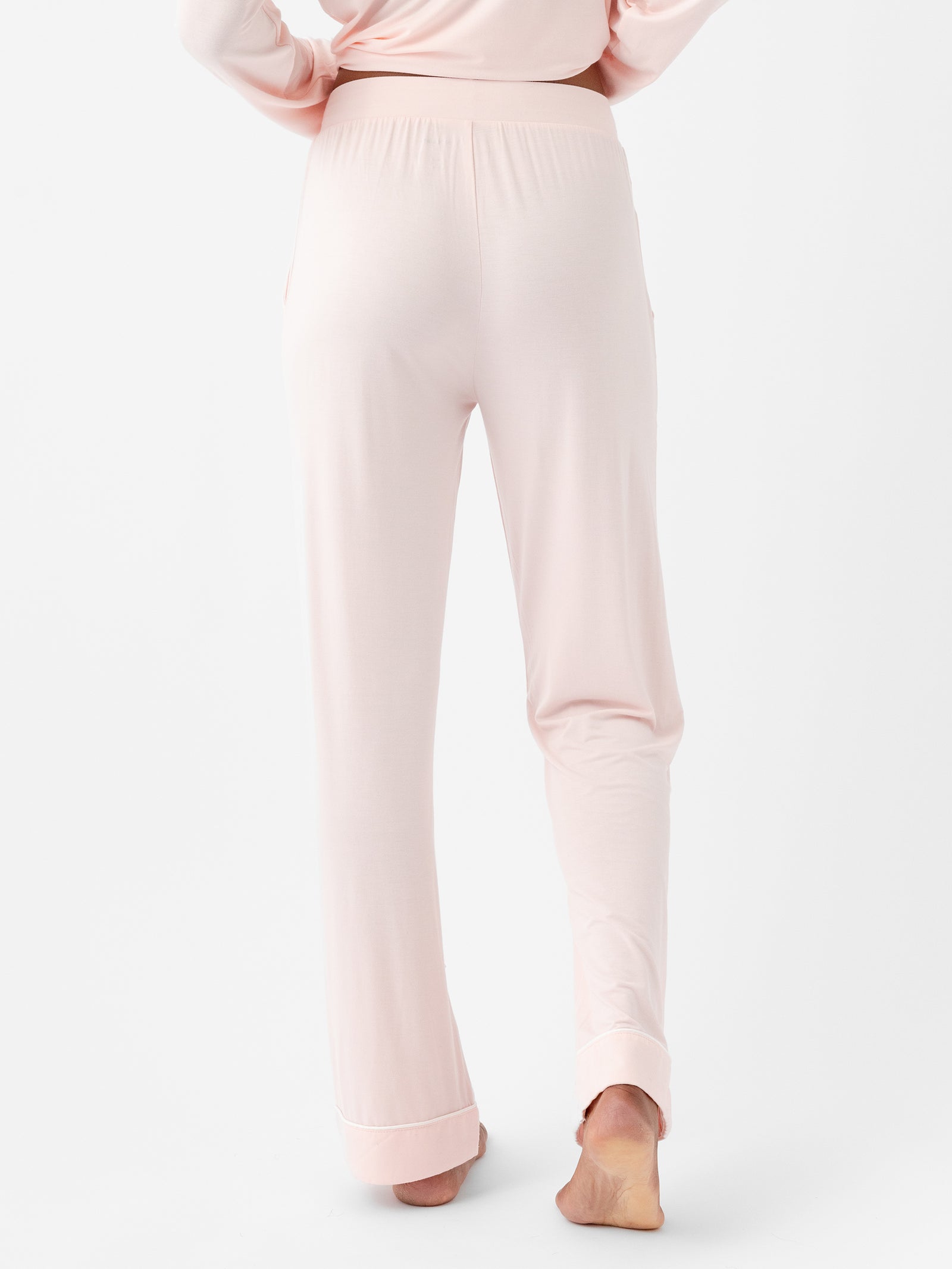 A person wearing blush pink Women's Stretch-Knit Bamboo Pajama Pants TALL by HIDE, shown from the back. The soft fabric pants feature an elastic waistband and gently drape over their legs. The person is barefoot, standing on their left foot with their right foot slightly lifted against a white background. 