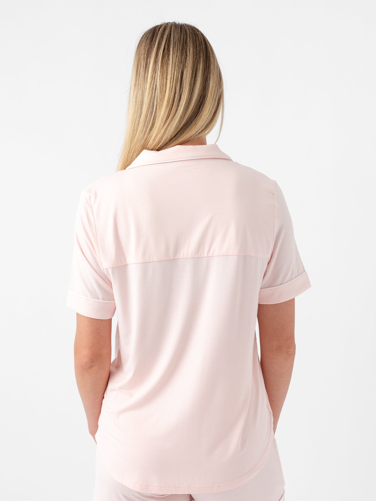 A person with long blonde hair is shown from the back, wearing a light pink Women's Short Sleeve Bamboo Pajama Top in Stretch-Knit by Cozy Earth, along with matching shorts. The background is plain white. |Color:Blossom