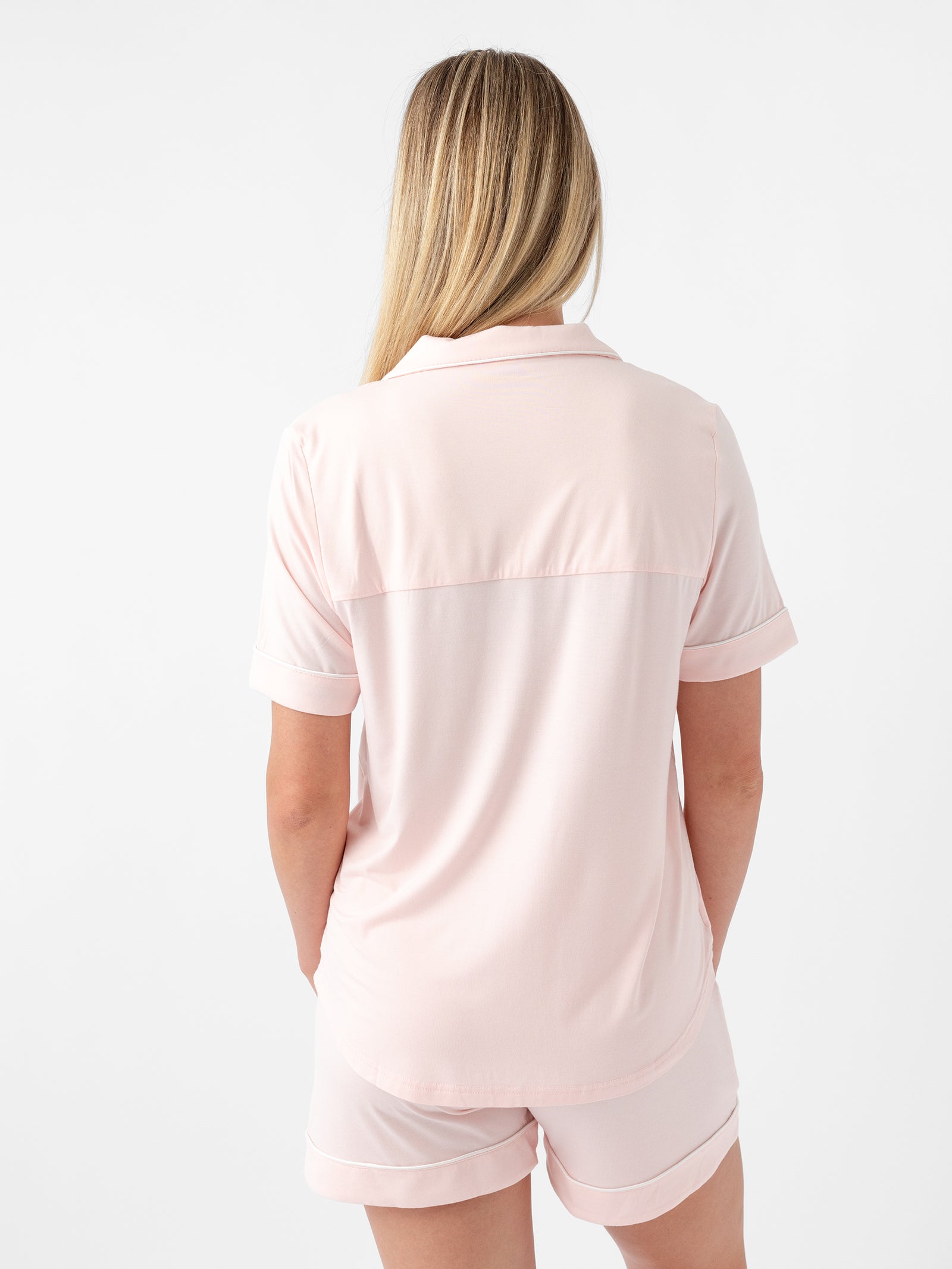 A person with long blonde hair is shown from the back, wearing a light pink Women's Short Sleeve Bamboo Pajama Top in Stretch-Knit by Cozy Earth, along with matching shorts. The background is plain white. 