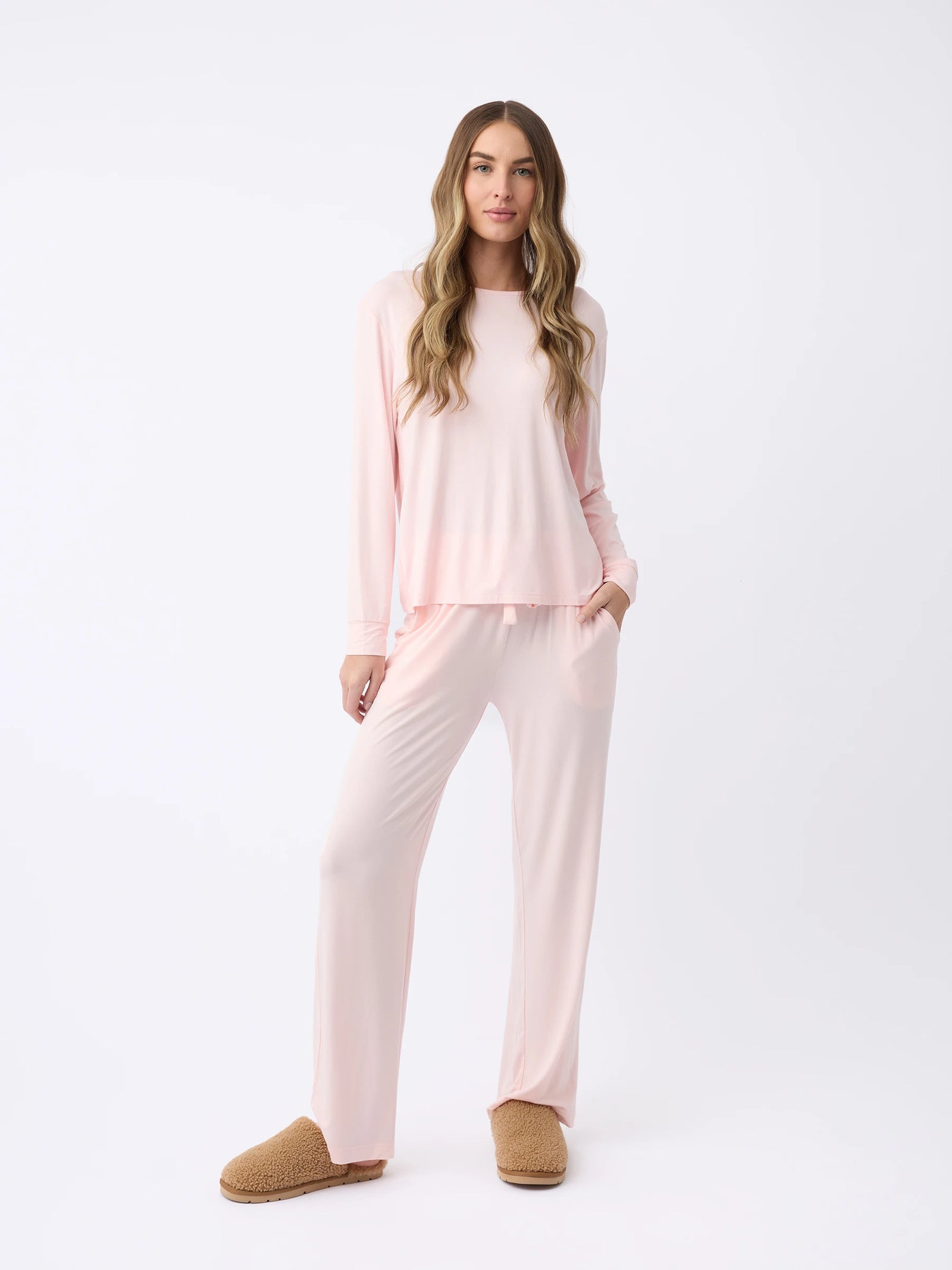 A woman with long, wavy hair stands relaxed against a white background wearing Cozy Earth's light pink Women's Stretch Knit Bamboo Long Sleeve Lounge Tee and brown slippers, with one hand in their pocket. 