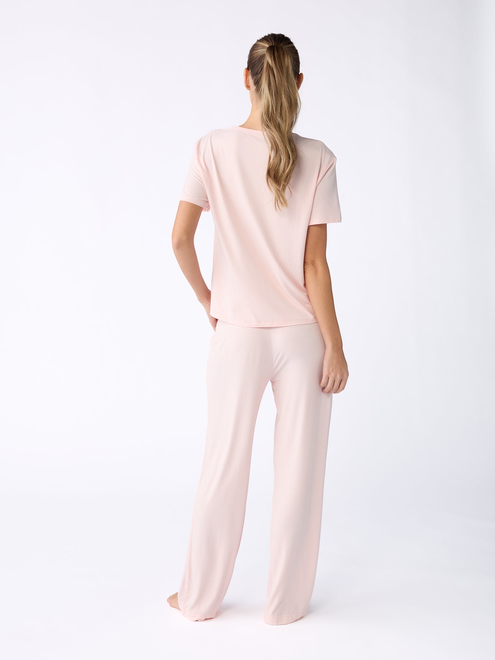 A person with long hair is standing barefoot, facing away from the camera. They are wearing a light pink pajama set featuring Cozy Earth's short-sleeved top and Stretch Knit Bamboo Pant, all set against a plain white background. 