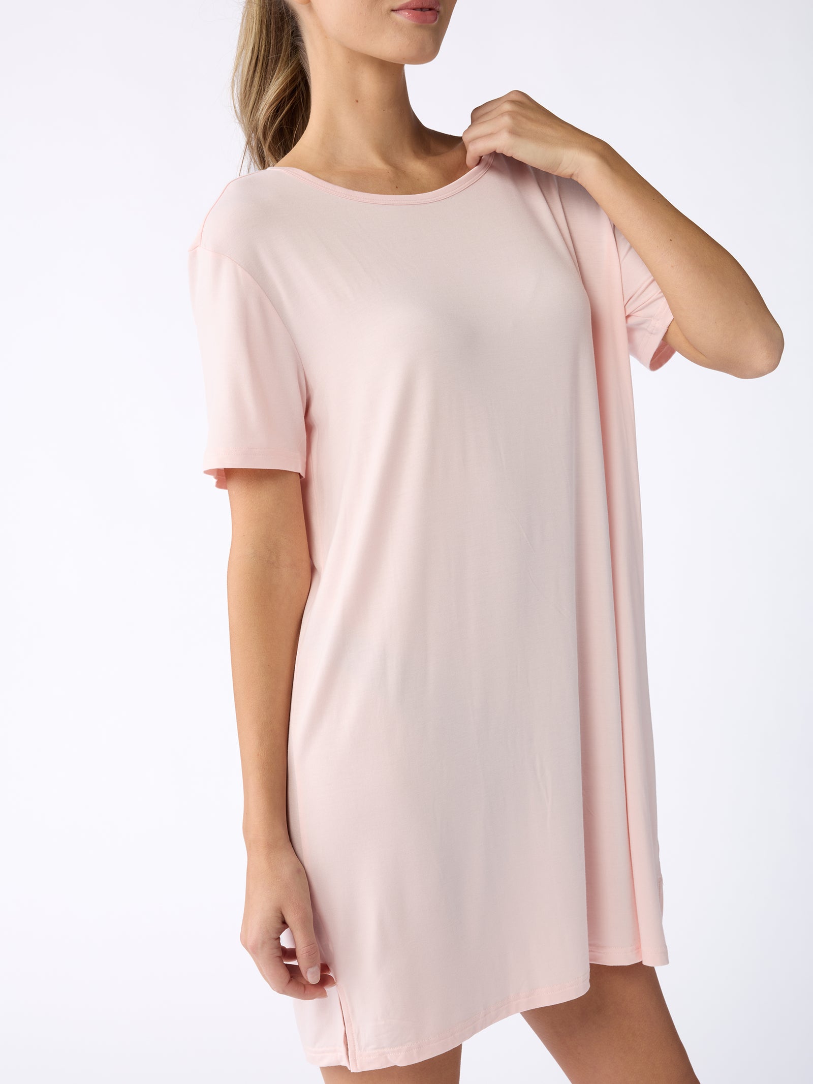 A woman is wearing the Women's Bamboo Stretch Knit Sleep Dress by Cozy Earth in a light pink hue. The dress features short sleeves, a loose fit, and a round neckline. She is partially turned to the side against a plain white background, with her long hair tied back in a ponytail. 