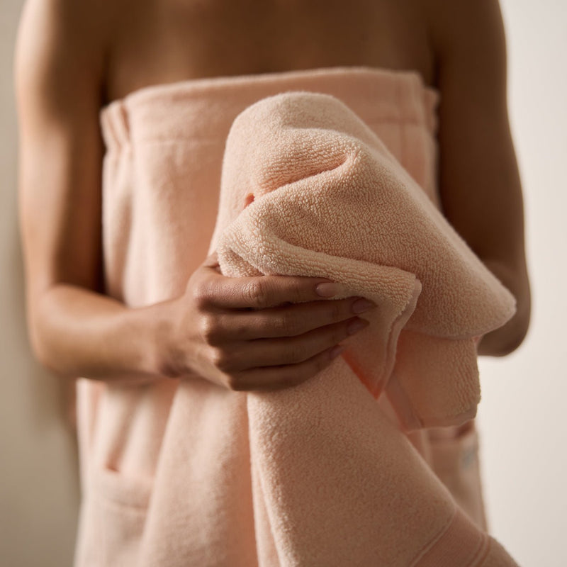 A person holds a soft, peach-colored Luxe Bath Sheet from Cozy Earth against their chest, while wearing another matching towel wrapped around their body. The background is neutral and out of focus.