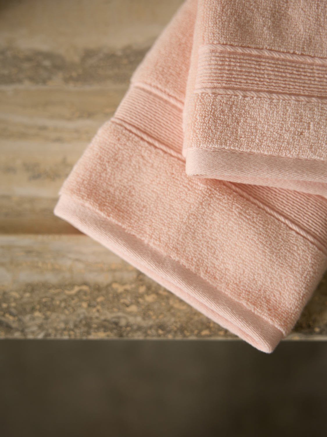 Two folded Luxe Hand Towels from Cozy Earth, in a peach color, rest on a textured wooden surface. Their soft texture and simple design are showcased against an earthy-toned background, complementing their gentle hue. 