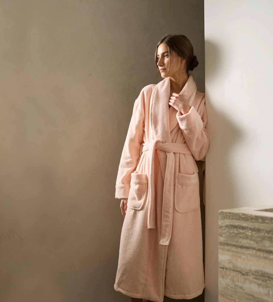 Woman in a pink bathrobe stands by a wall, gazing to the side. Her hair is tied back, and she looks relaxed. The setting is softly lit, creating a serene atmosphere.