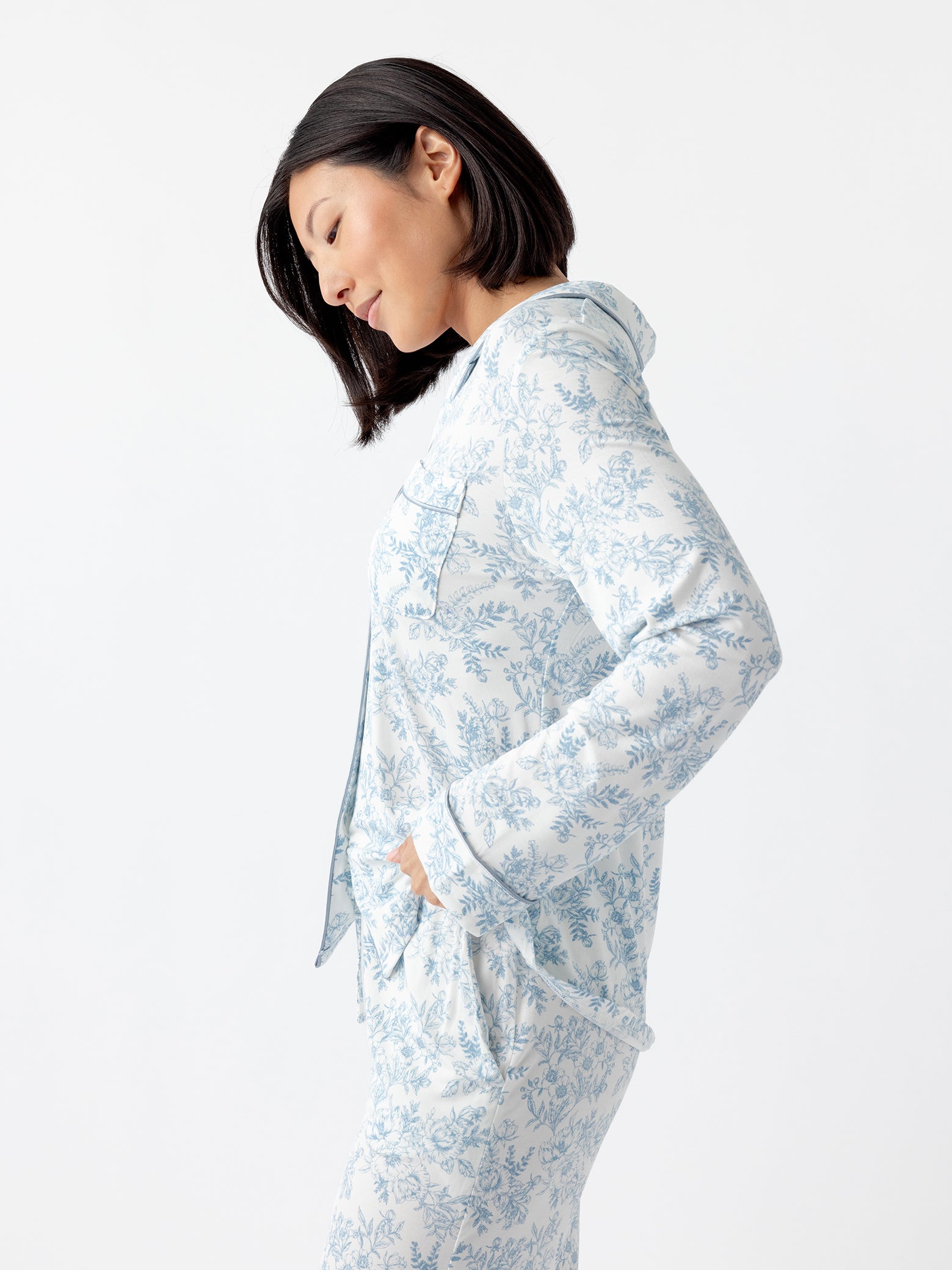 A woman with shoulder-length dark hair stands sideways with hands in pockets, wearing Cozy Earth Women's Bamboo Stretch-Knit Long Sleeve Pajama Top in light blue floral-patterned design featuring a hoodie, against a white backdrop. 