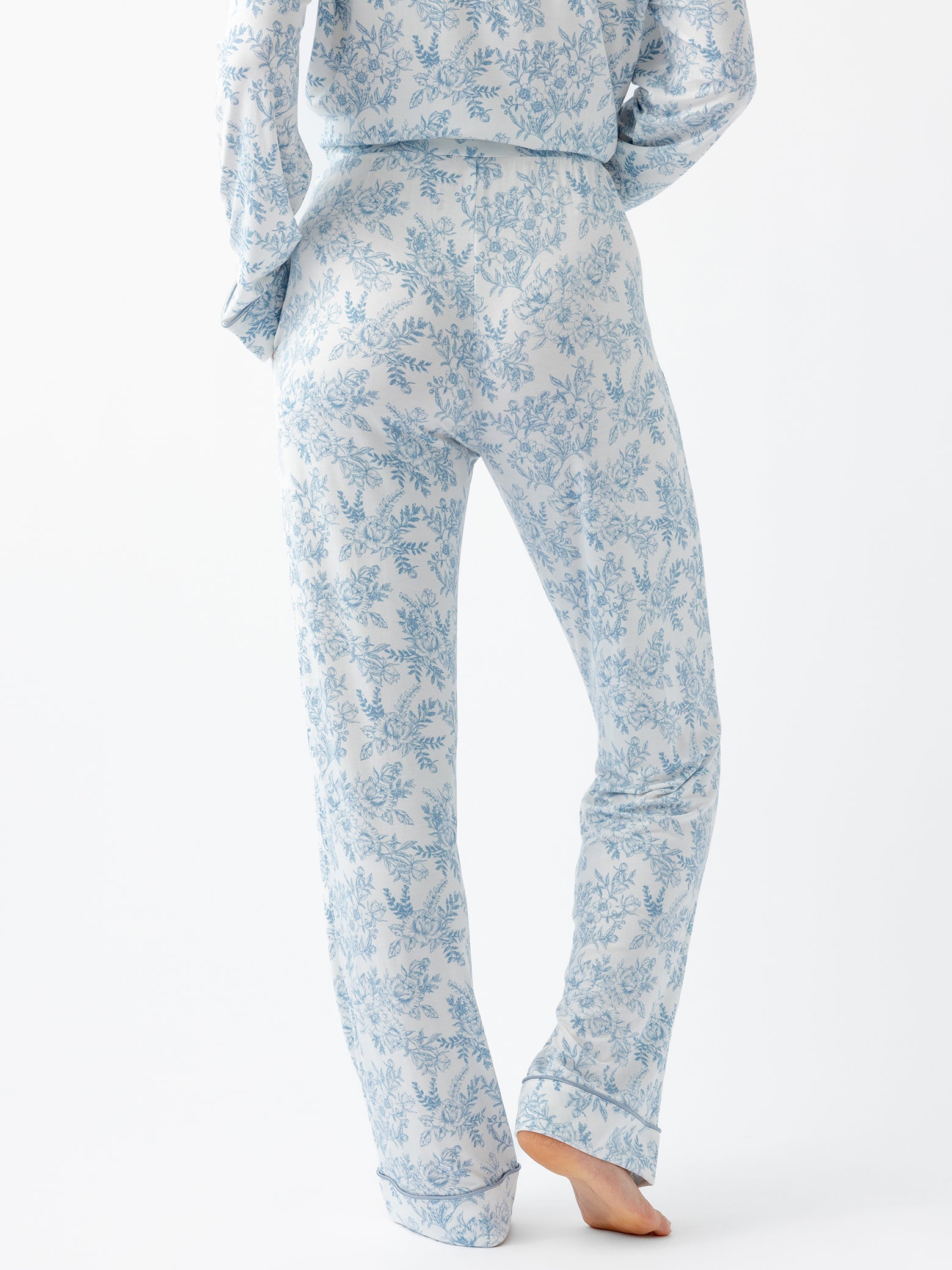 A person is shown from the back, barefoot on a light surface, wearing Cozy Earth Women's Bamboo Stretch-Knit Pajama Pant in a light blue and white floral pattern with long sleeves. 