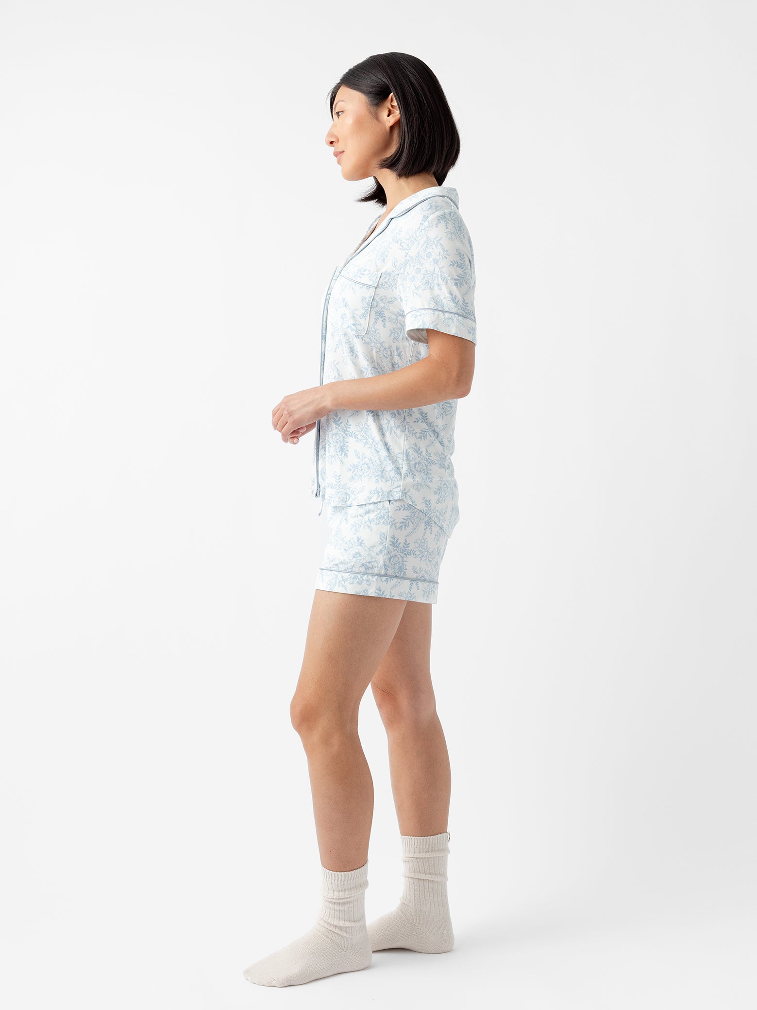 A person with short dark hair stands in profile wearing the Women's Bamboo Stretch-Knit Short Sleeve Pajama Top by Cozy Earth, in a light blue and white pattern, paired with white socks against a plain white background. 