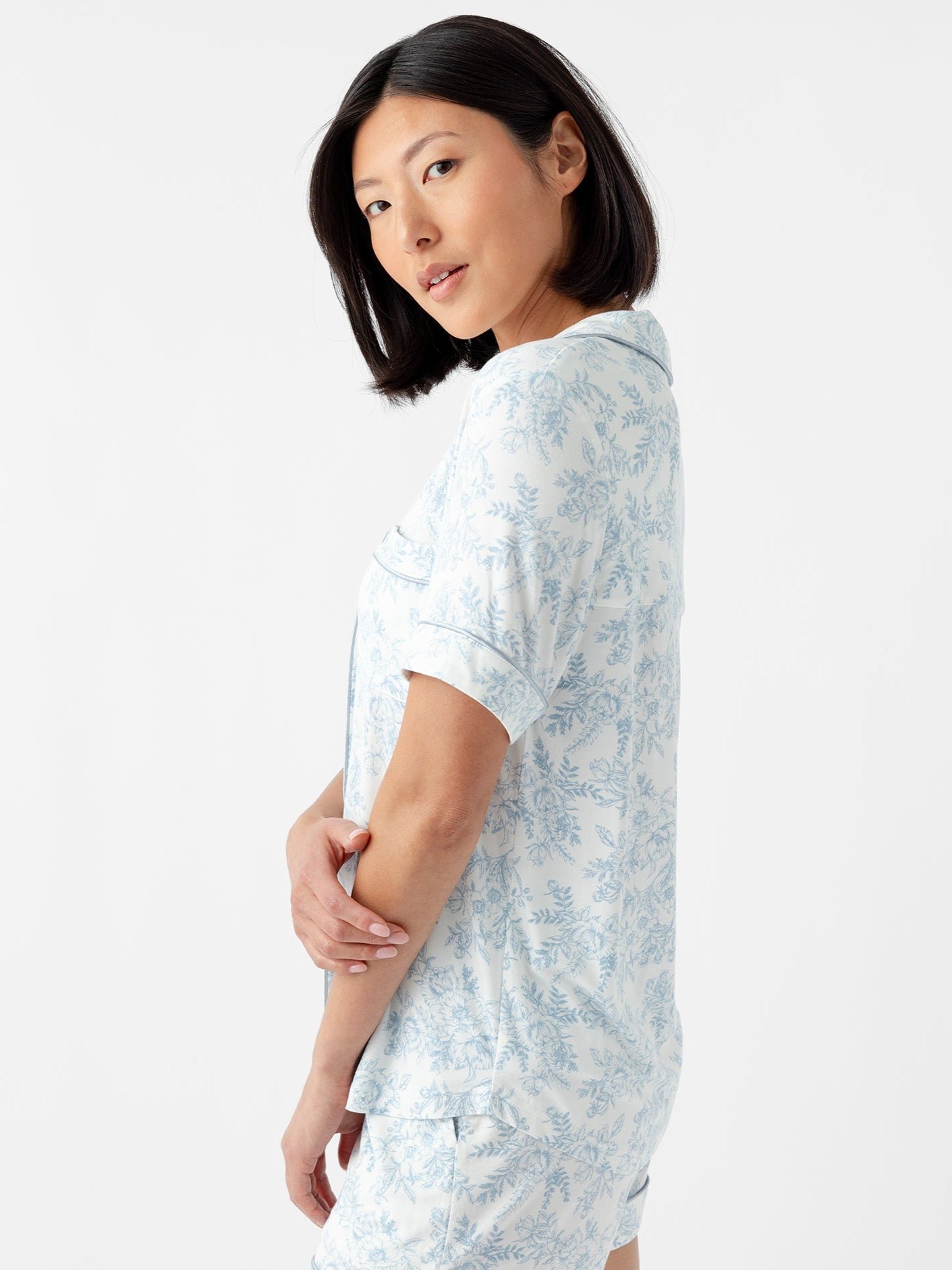 A woman with shoulder-length black hair wears a Women's Bamboo Stretch-Knit Short Sleeve Pajama Top by Cozy Earth in light blue floral pattern. She stands sideways, gazing over her shoulder against a plain white background. 