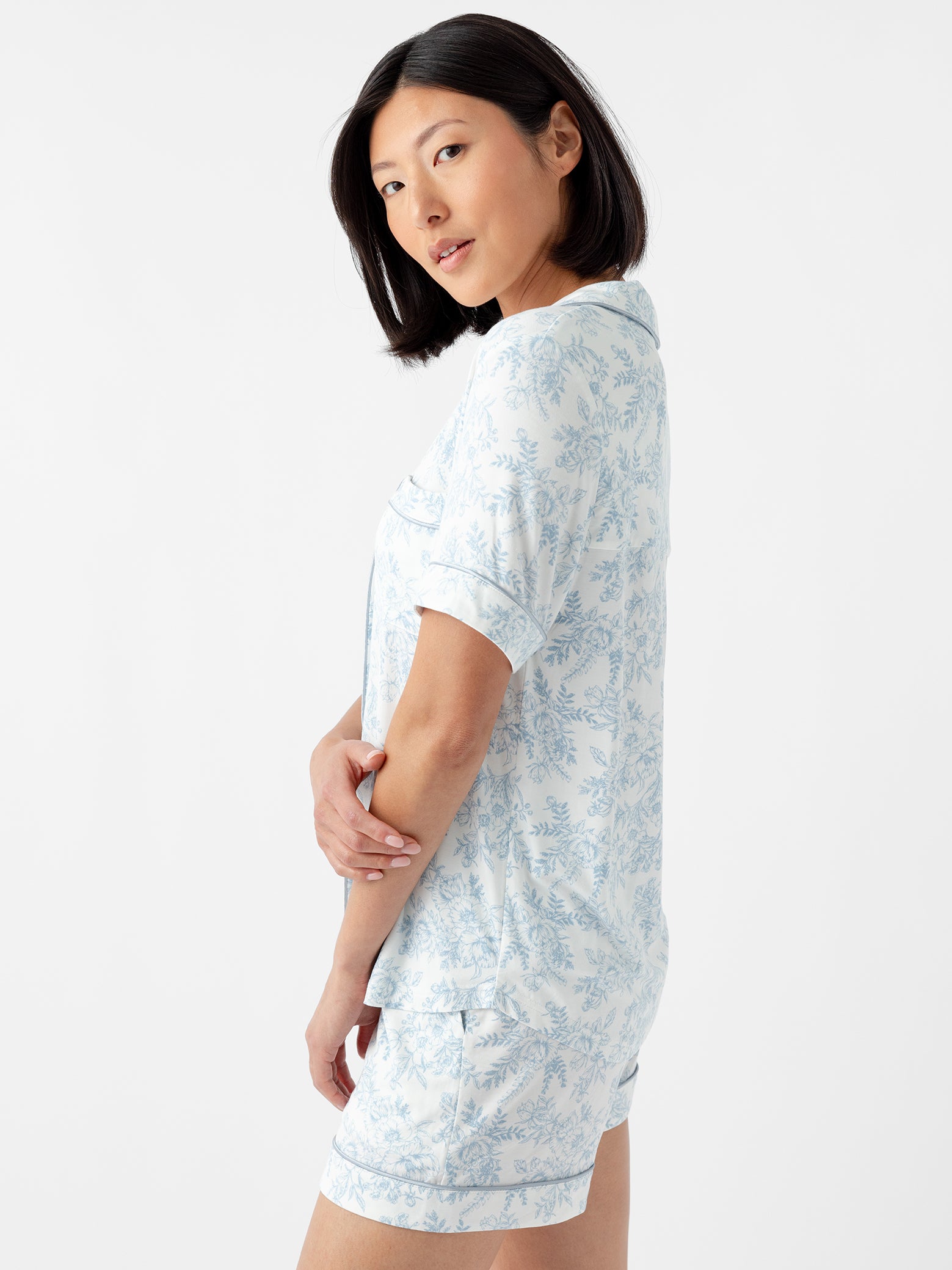 A woman with shoulder-length black hair wears a Women's Bamboo Stretch-Knit Short Sleeve Pajama Top by Cozy Earth in light blue floral pattern. She stands sideways, gazing over her shoulder against a plain white background. 