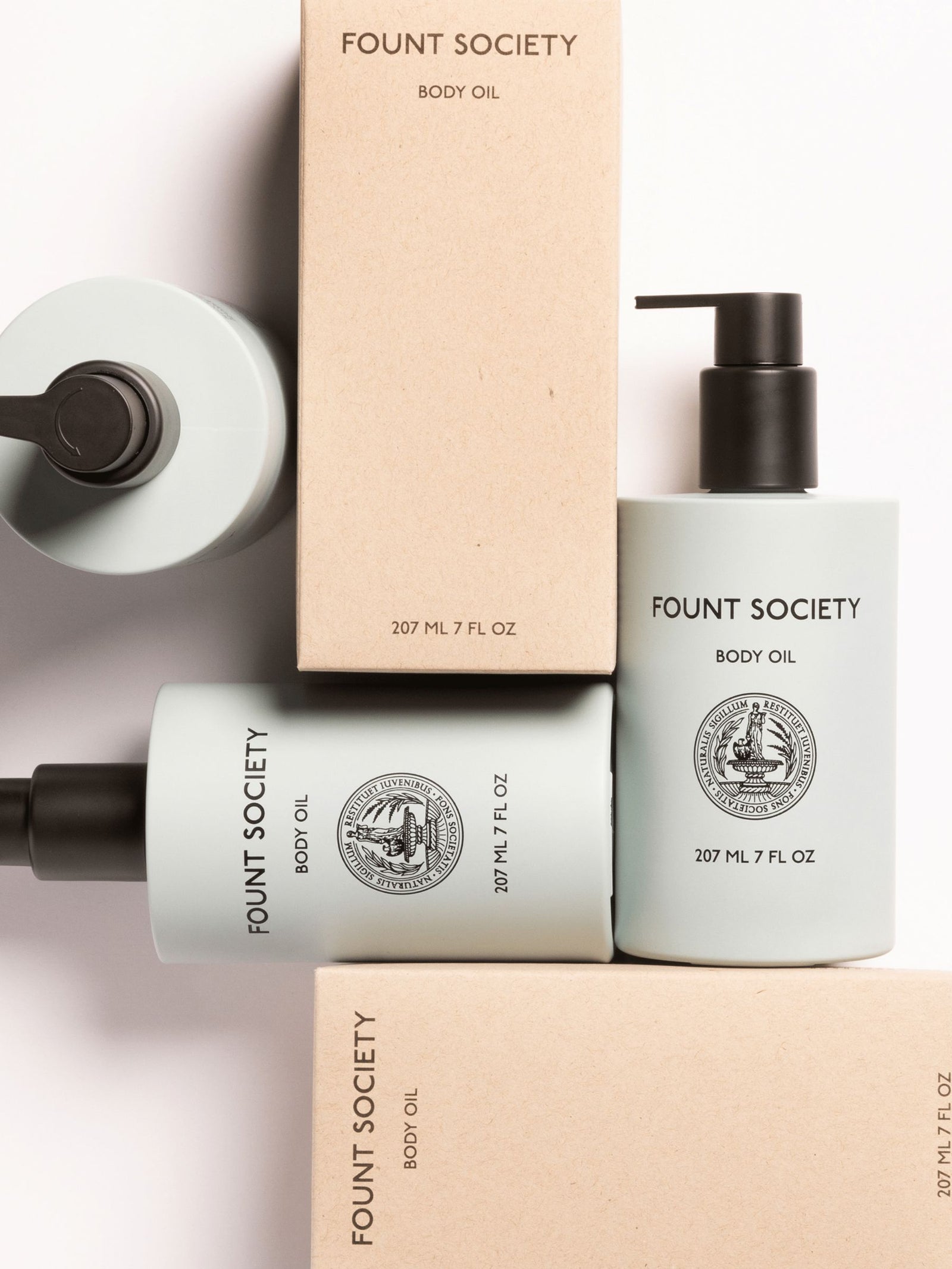 A flat lay features Cozy Earth Body Oil bottles and boxes: two light green pump bottles and two beige boxes with product labeling are aesthetically arranged on a white surface.